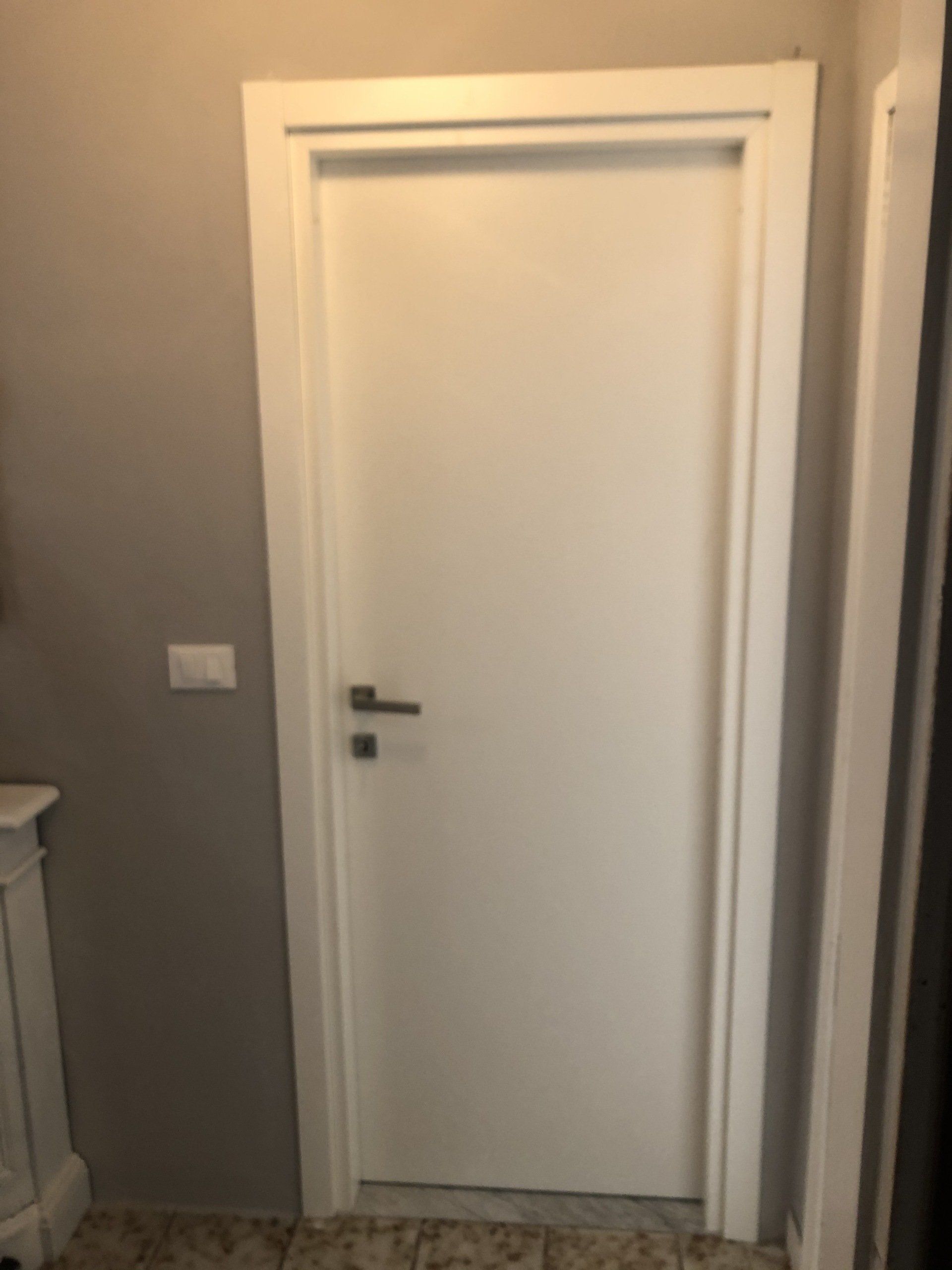 Porta bianca con maniglia e telaio argentati, contro una parete grigia.
