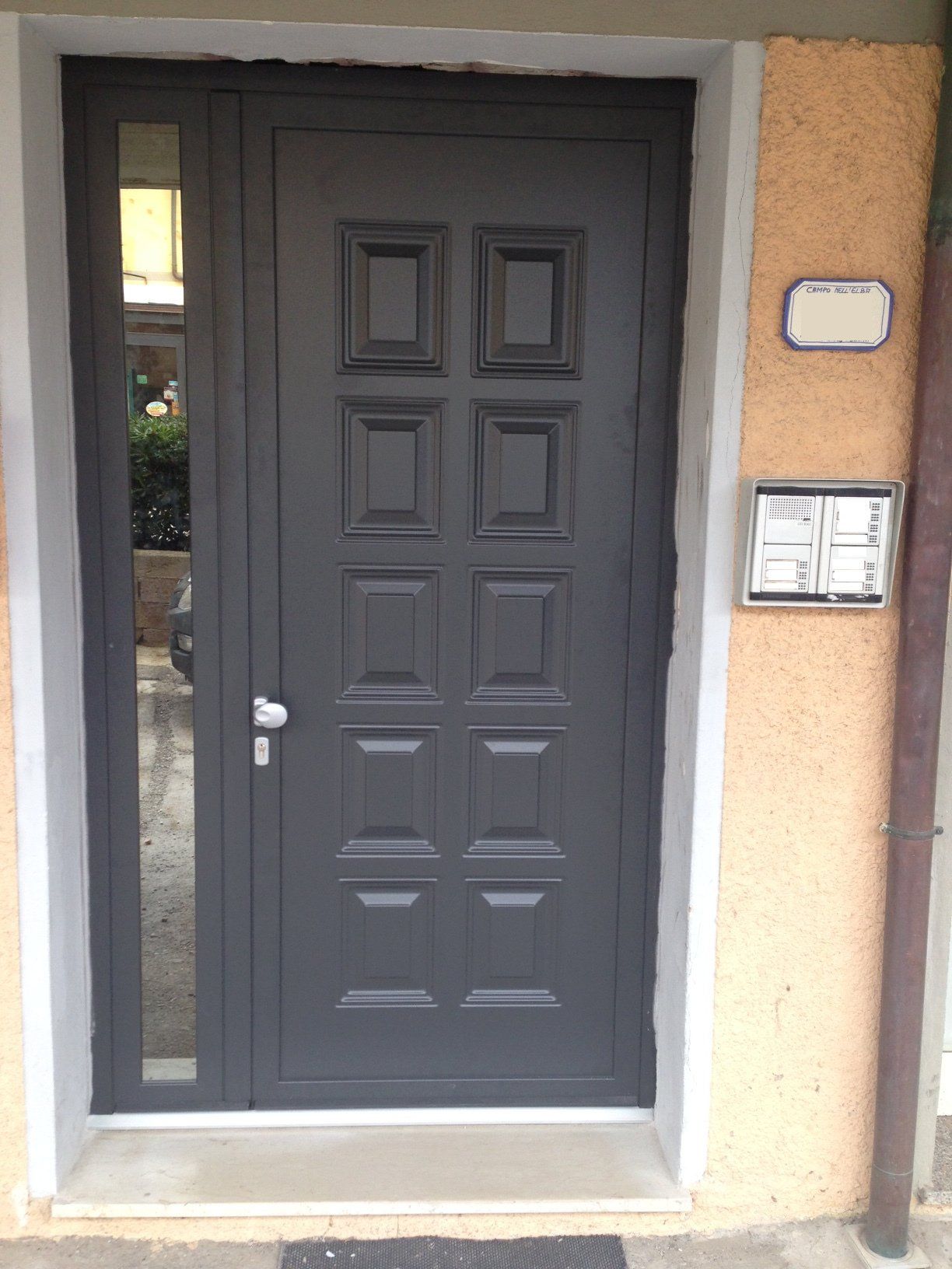 Porta d'ingresso grigio scuro con pannelli rettangolari e una finestra laterale in vetro, inserita in una parete esterna di colore chiaro.