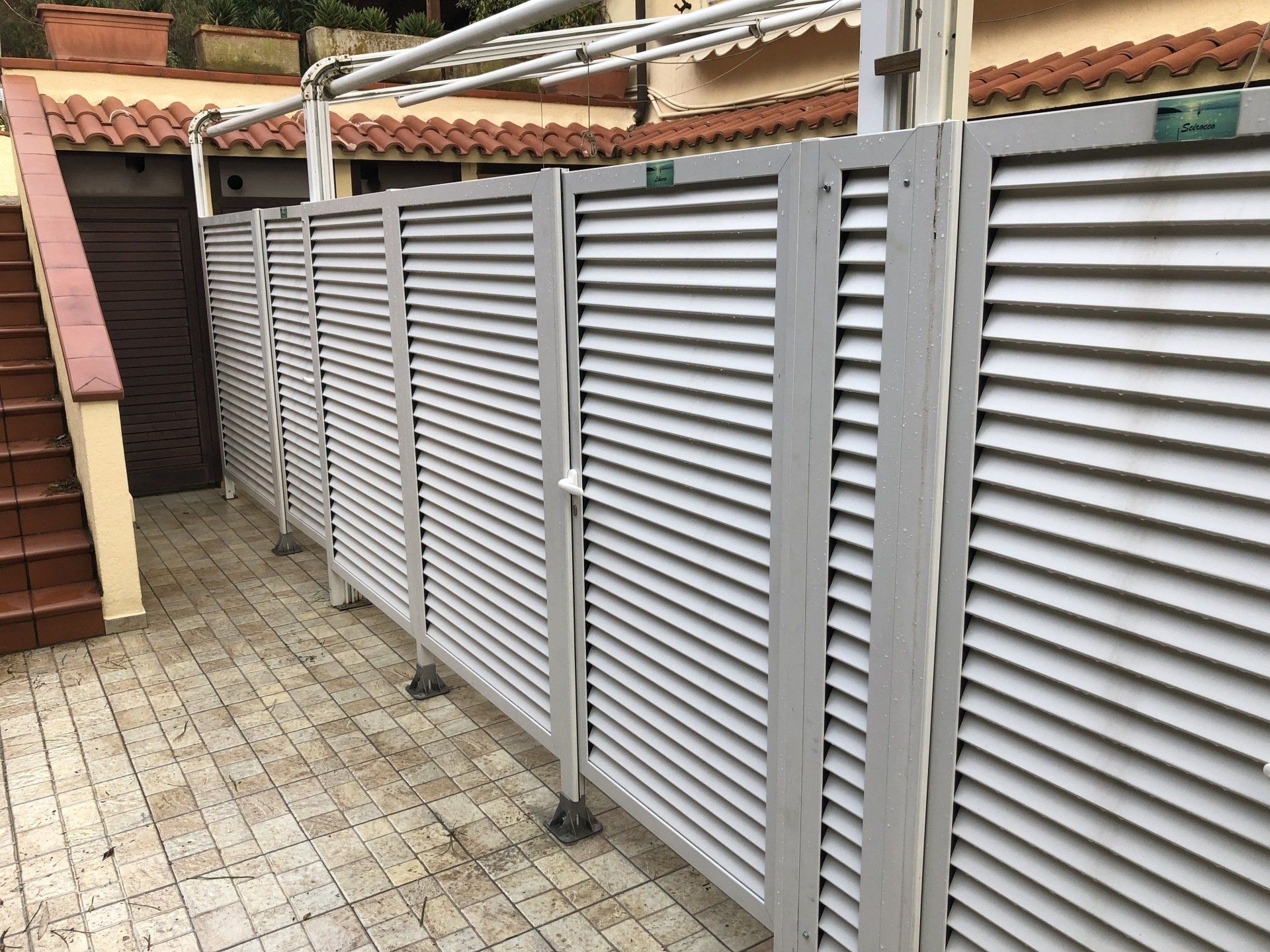 Fila di schermature per la privacy esterne con persiane grigie, posizionate su un patio piastrellato.