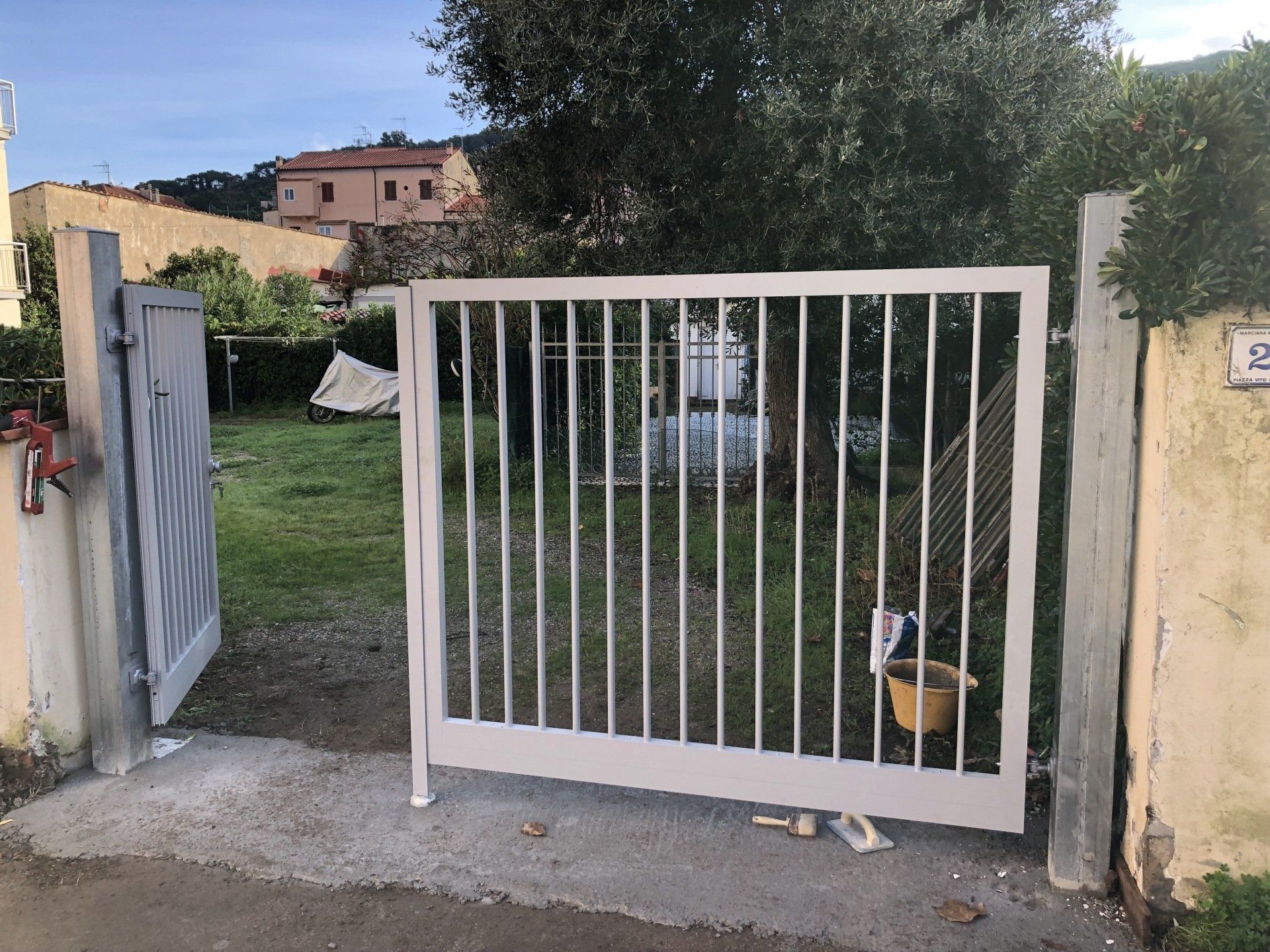 Un cancello di metallo grigio chiaro, parzialmente aperto, con sbarre verticali. Un secondo cancello è aperto sulla sinistra.