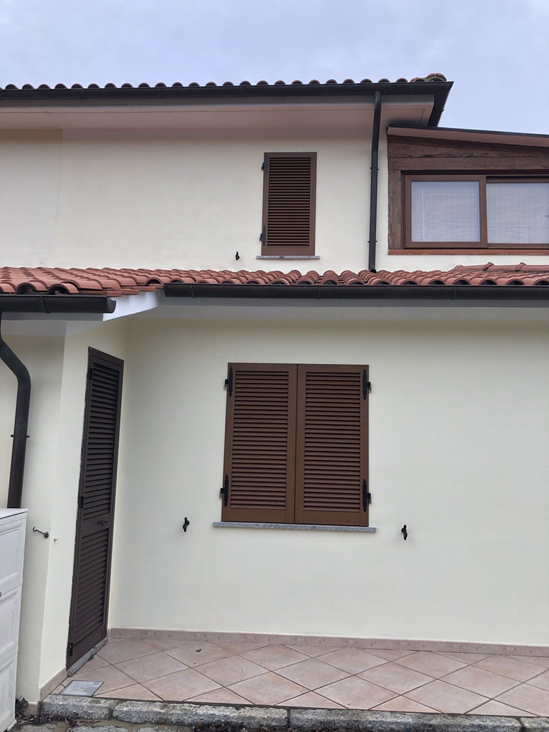 Casa beige con persiane marroni, patio piastrellato e tetto in tegole di terracotta.