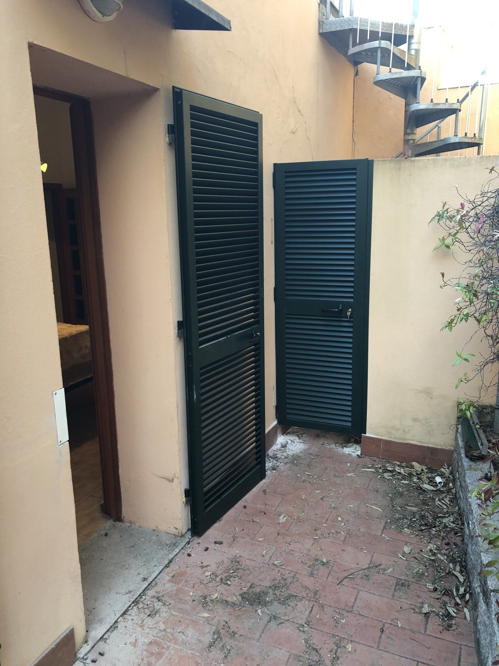 Due porte a persiana verde scuro si aprono su un patio in mattoni. È visibile una scala a chiocciola.
