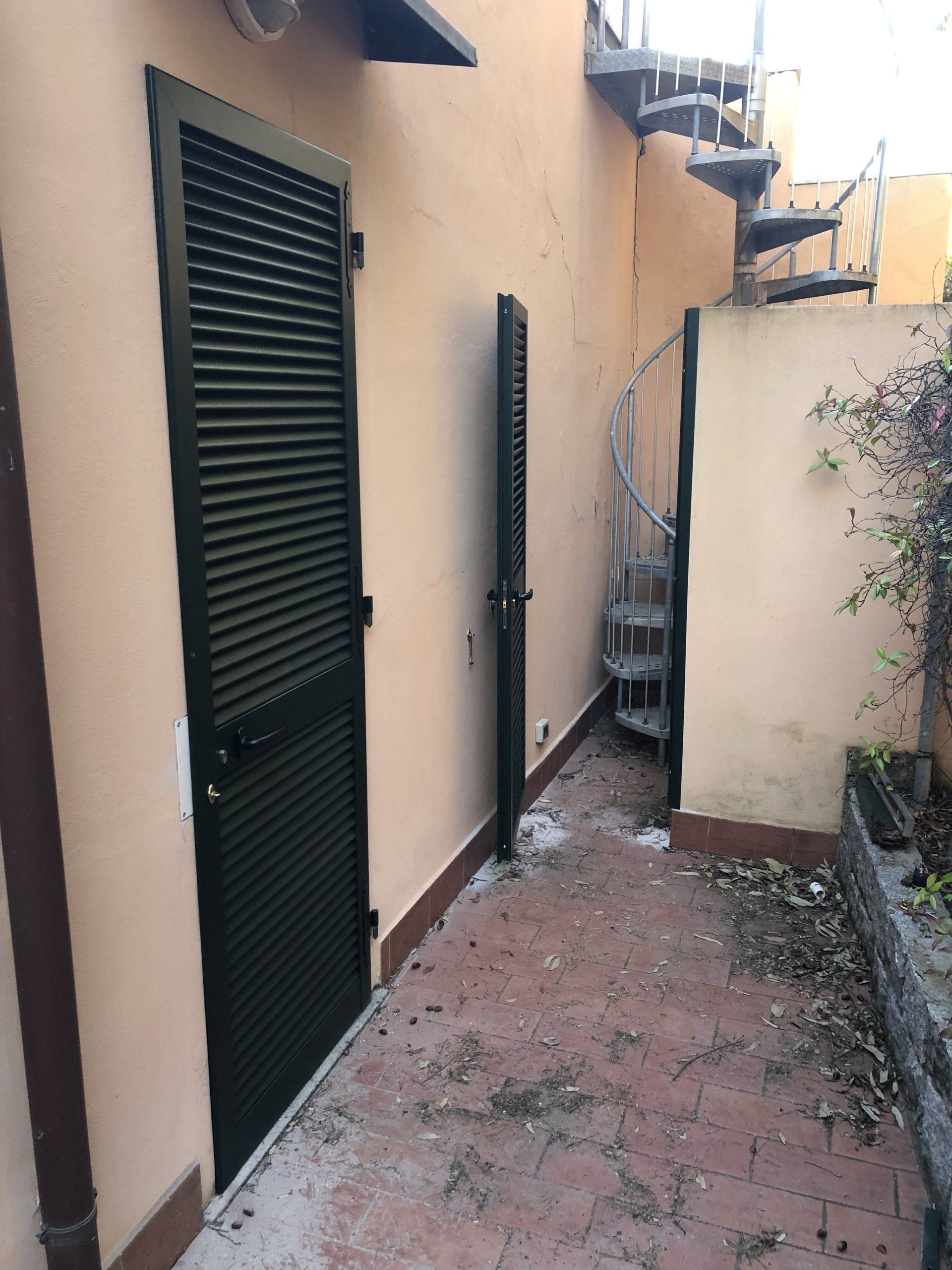 Uno spazio esterno stretto con una porta scura con persiane, un cancello di metallo e una scala a chiocciola.