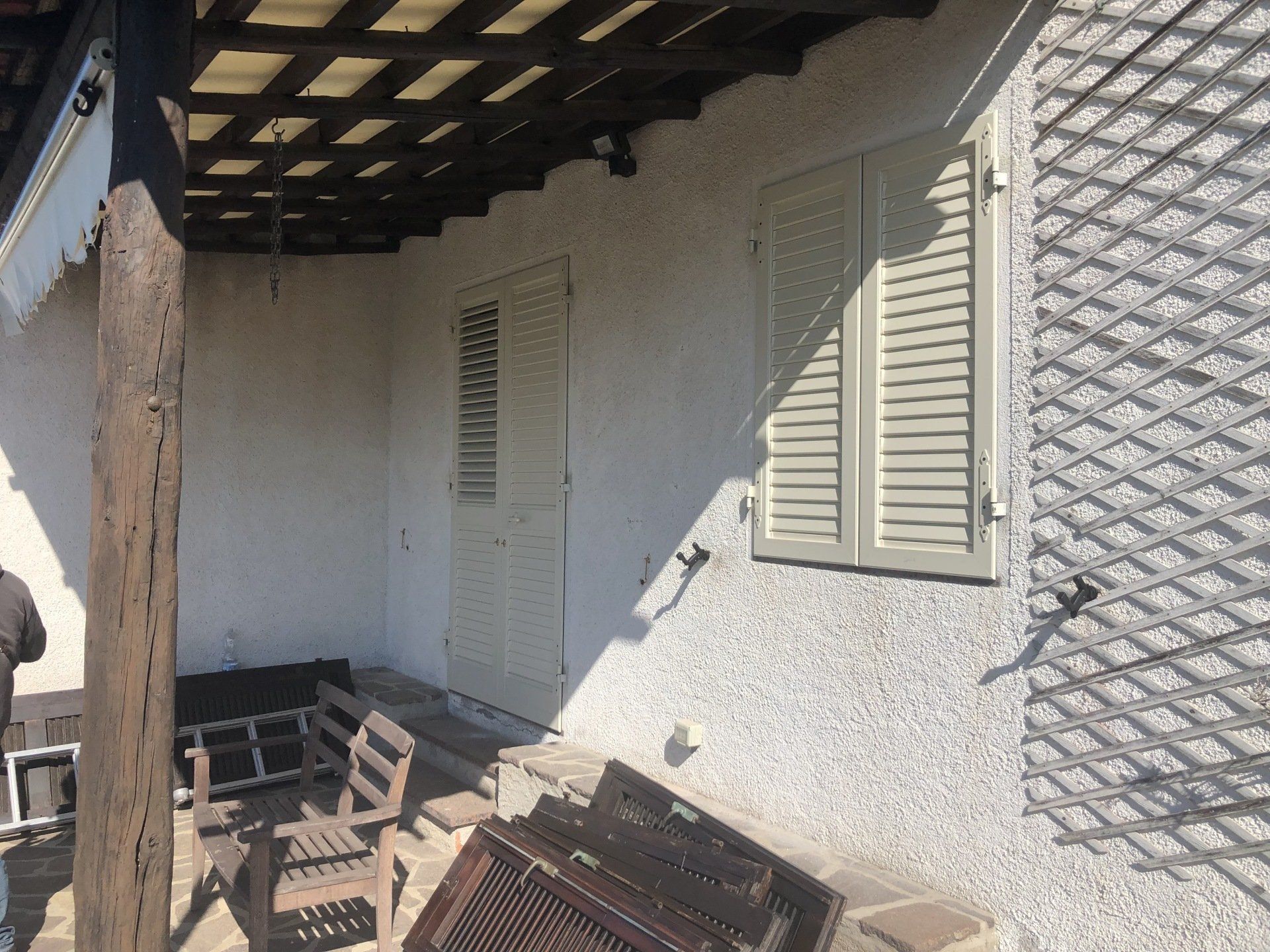 Esterno di un edificio con persiane chiuse di colore chiaro e un portico coperto da un pergolato.