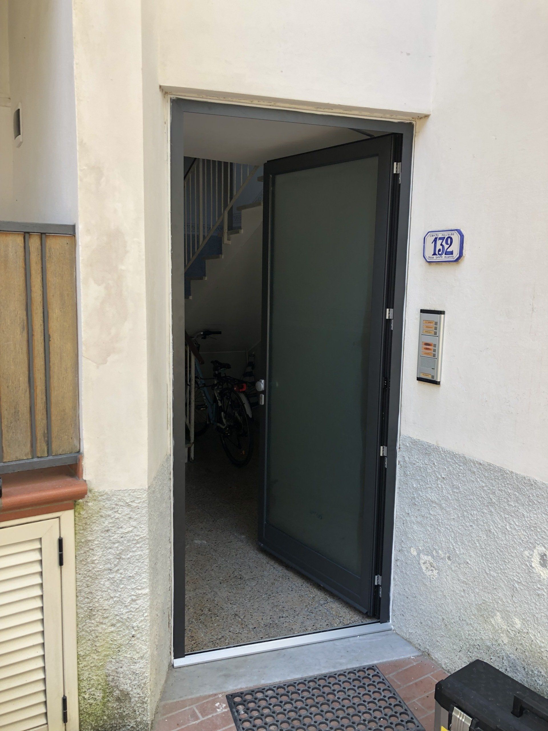 Porta aperta che dà su un edificio; porta in vetro smerigliato con cornice scura, scale visibili, bicicletta parcheggiata all'interno.