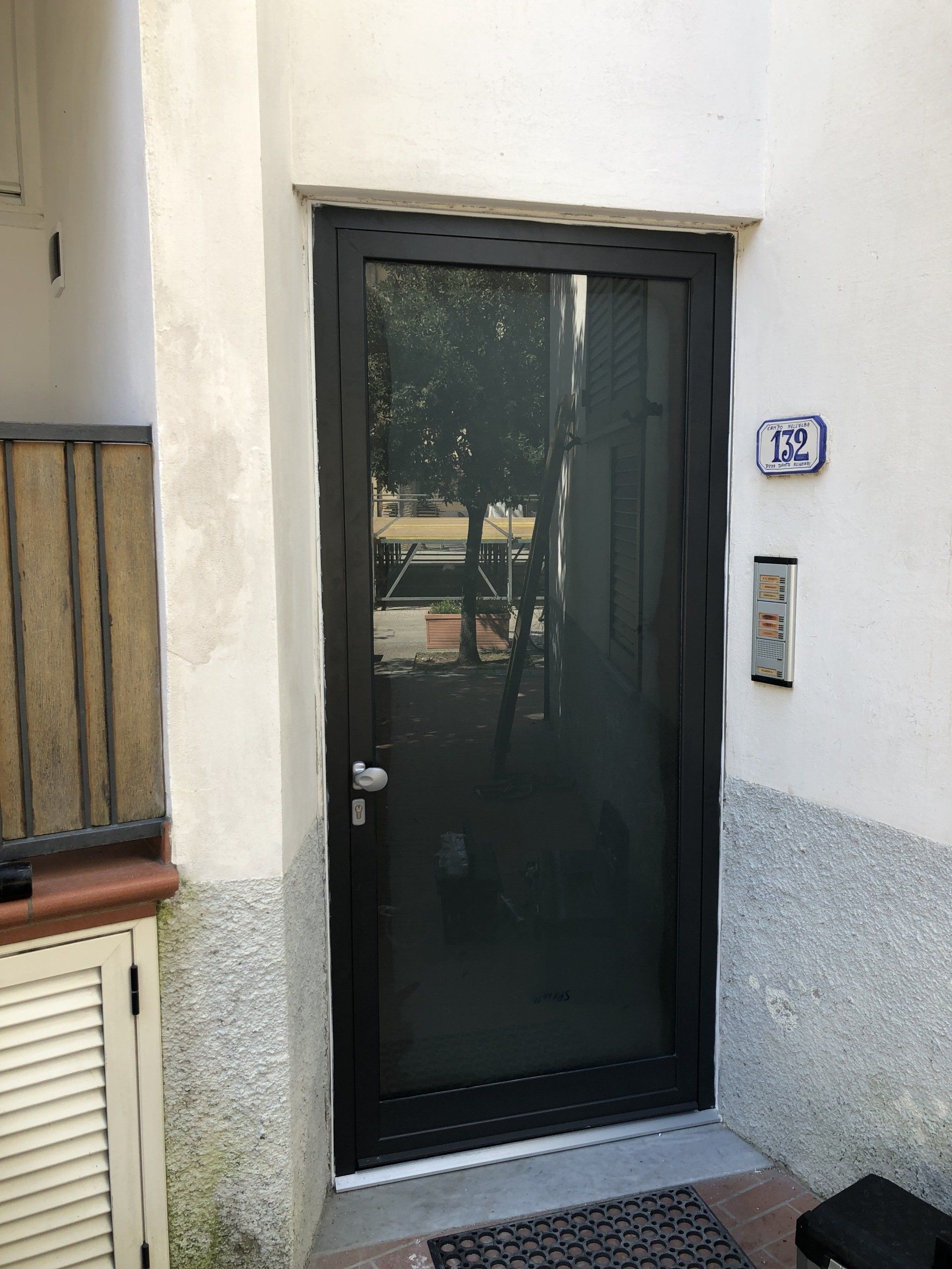 Porta nera con pannello in vetro, accanto a pareti bianche e grigie, campanello e targa con l'indirizzo visibili.