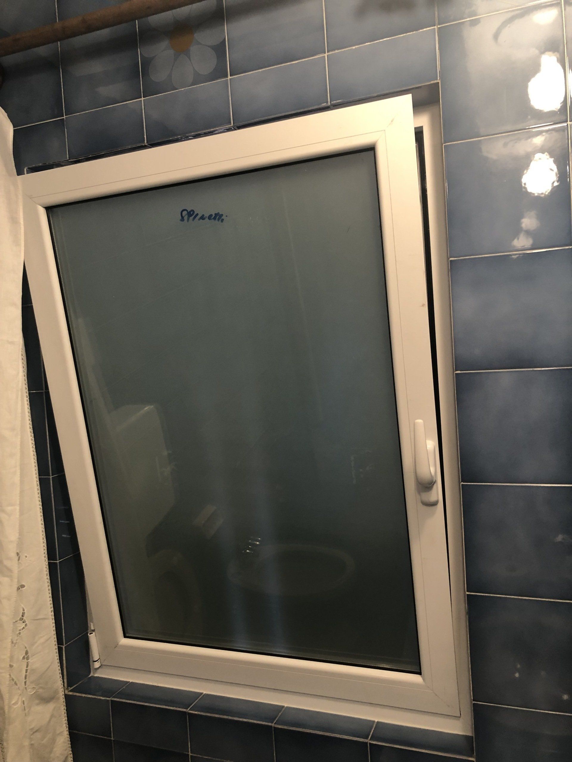 Finestra con cornice bianca in un bagno piastrellato di blu, leggermente socchiusa, con vetro smerigliato.