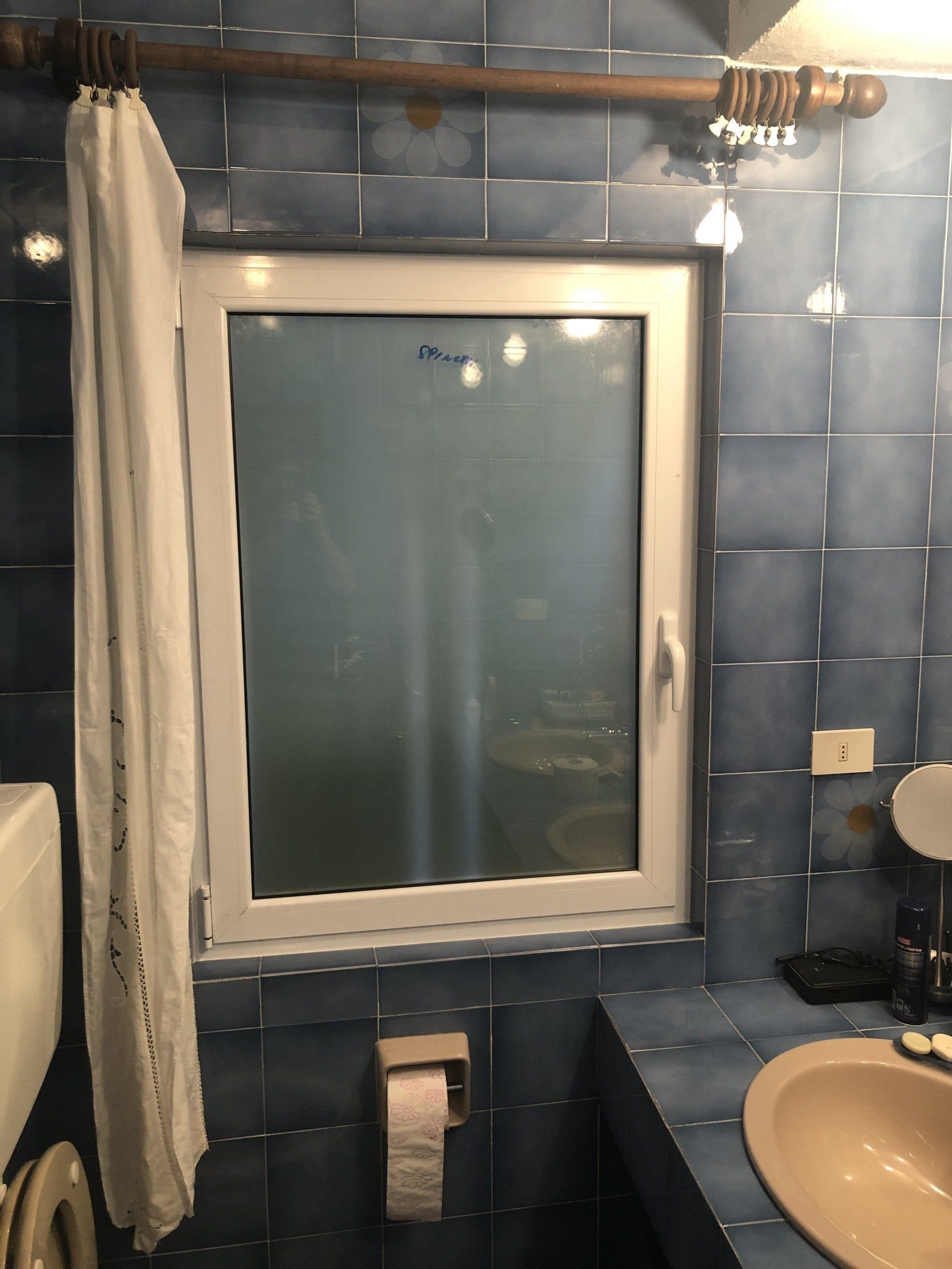 Bagno con pareti piastrellate blu, una finestra con vetro smerigliato e una tenda bianca.