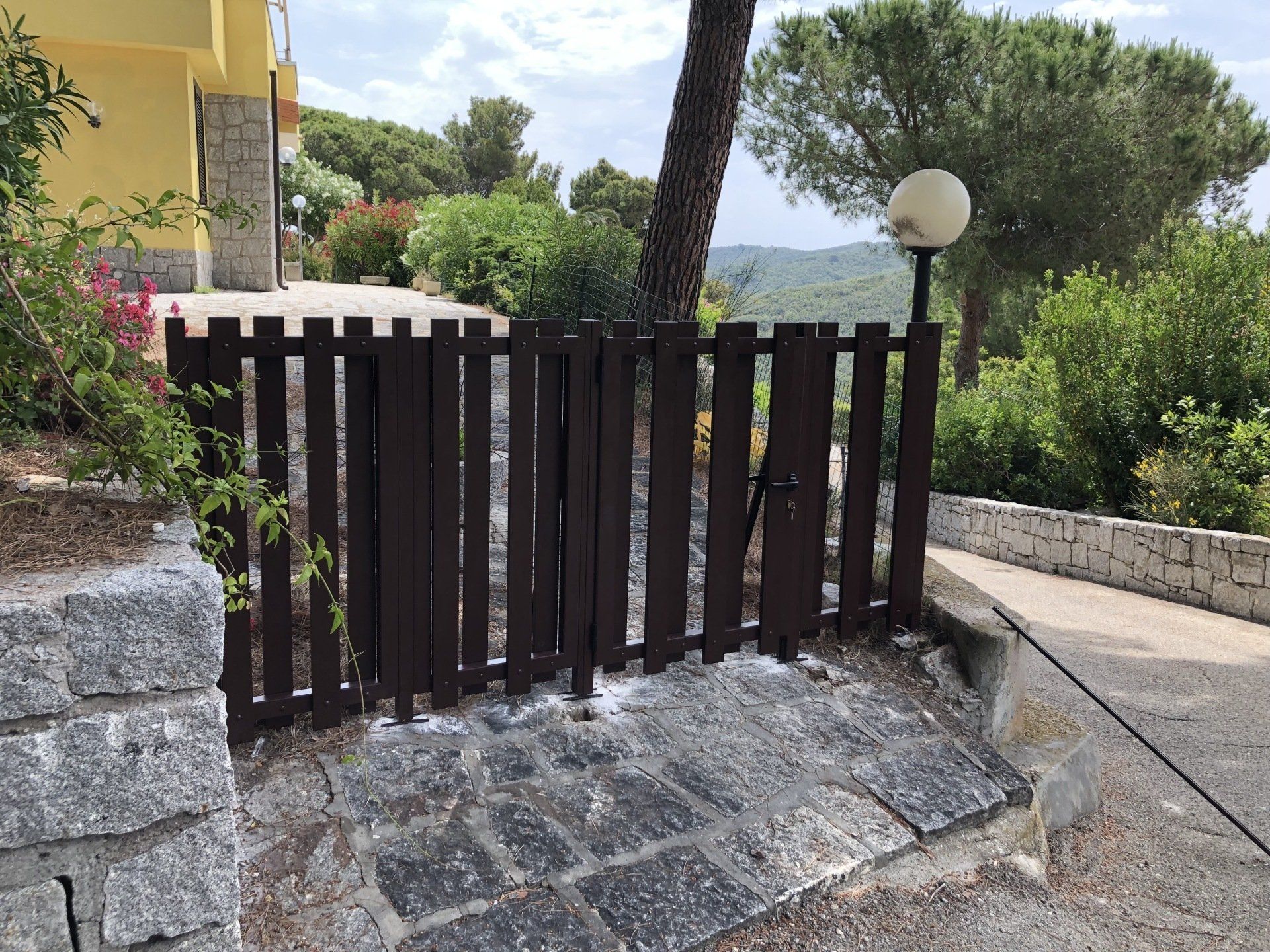 Cancello di legno marrone su un sentiero di pietra, che conduce a una casa con vegetazione sullo sfondo.