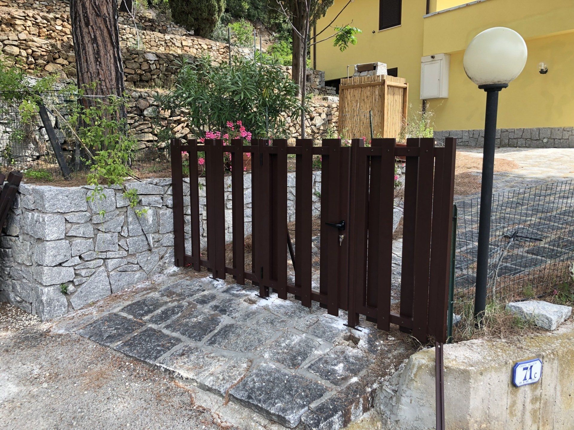 Cancello in legno marrone su gradini in pietra con un muro in pietra, un edificio giallo sullo sfondo e un lampione.