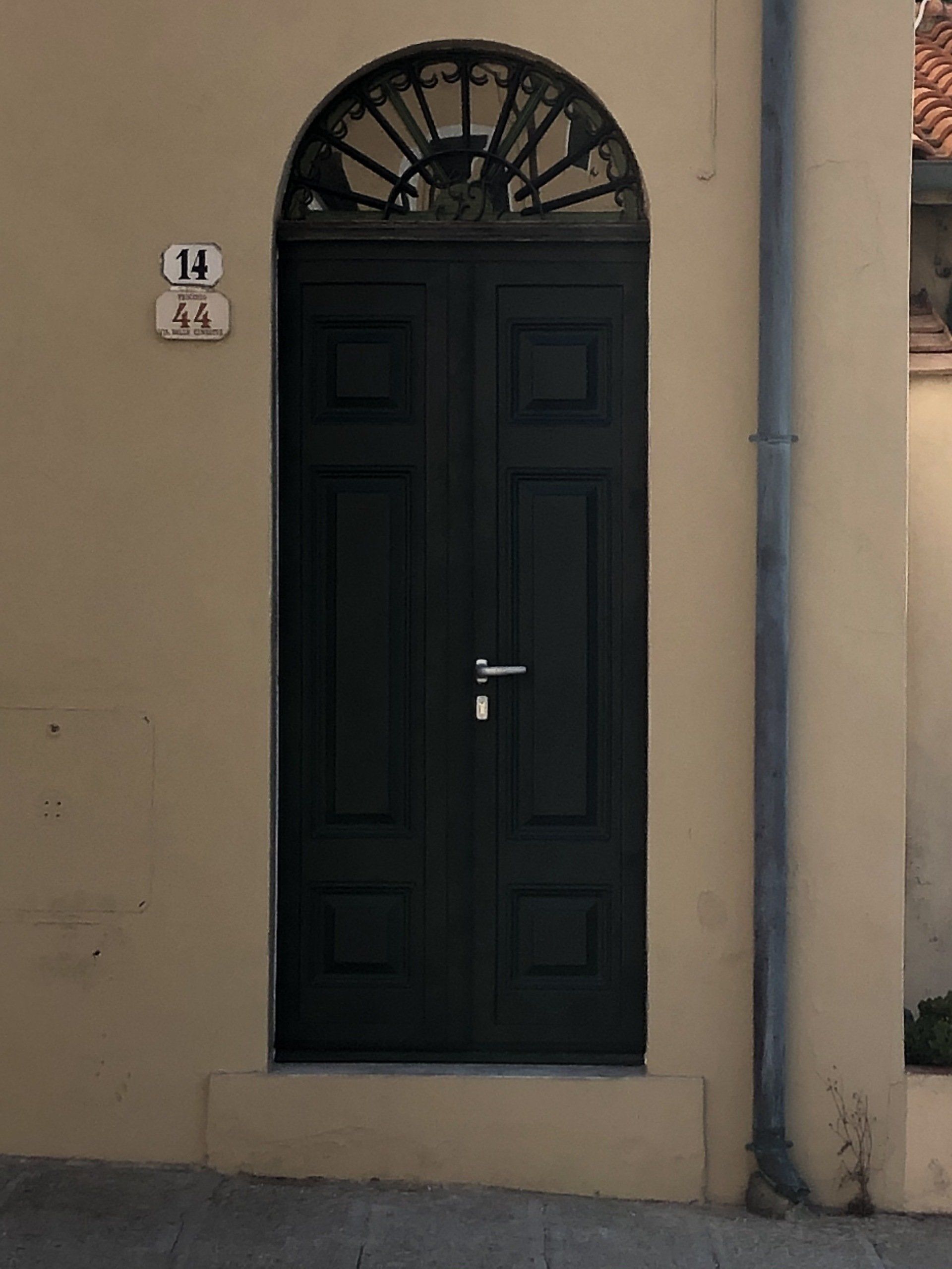 Porta ad arco nera con lavorazione in metallo, targa con indirizzo sulla parete beige e grondaia.