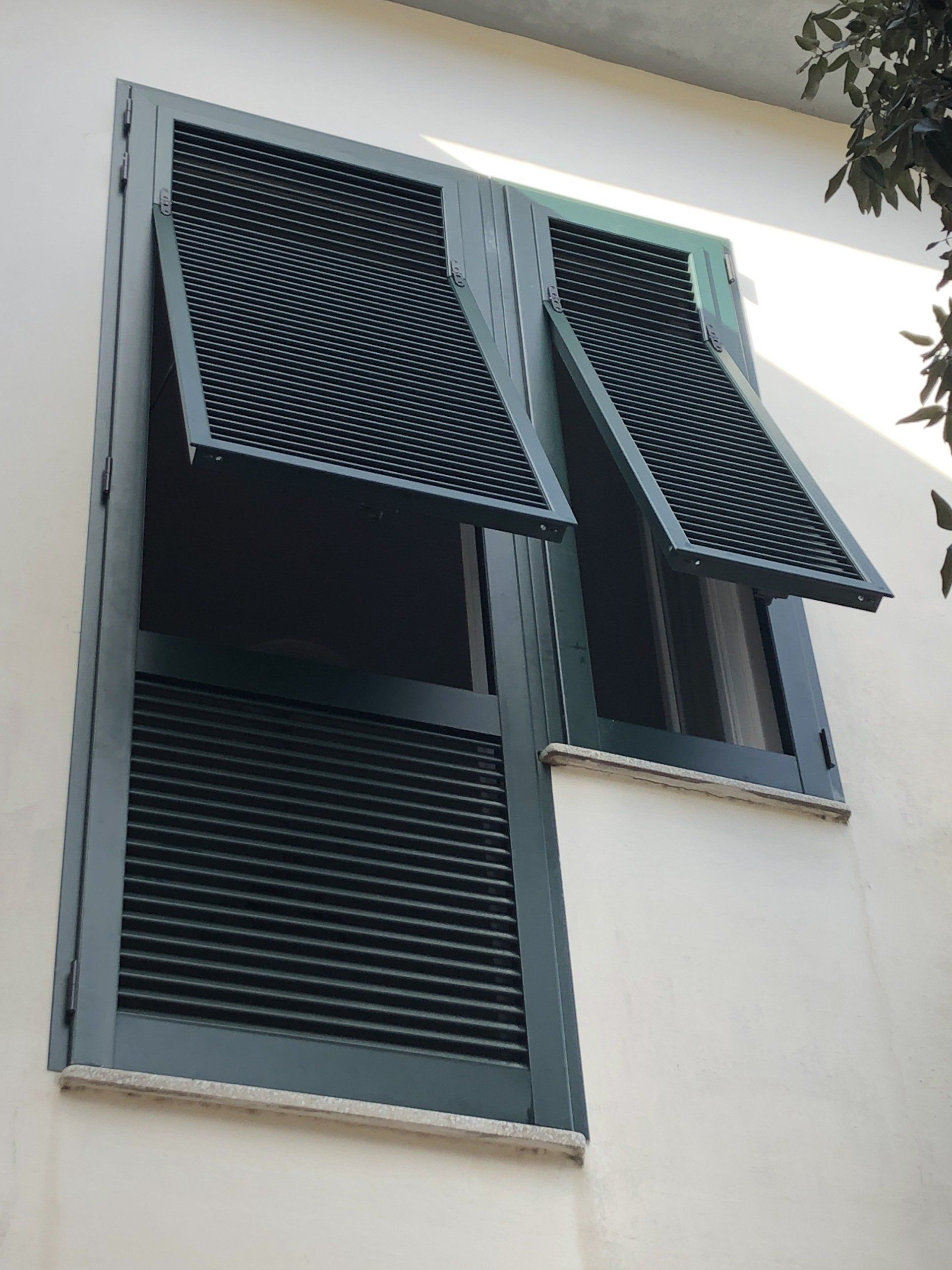 Finestra con persiane verde scuro su un edificio bianco; le persiane sono aperte, rivelando la finestra dietro di esse.