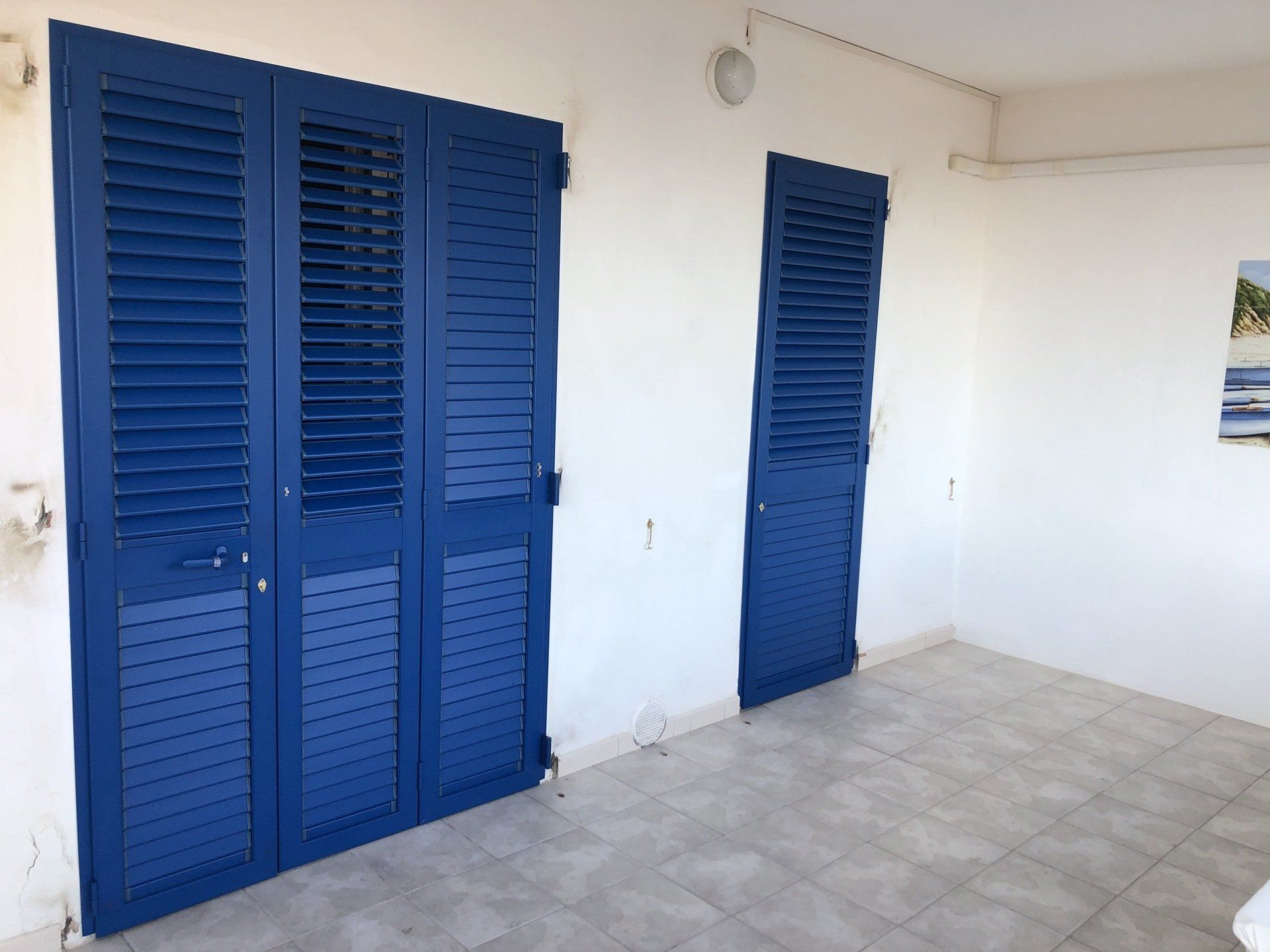 Persiane blu su una parete bianca, con porta coordinata. Una terrazza luminosa e soleggiata con pavimento in piastrelle.