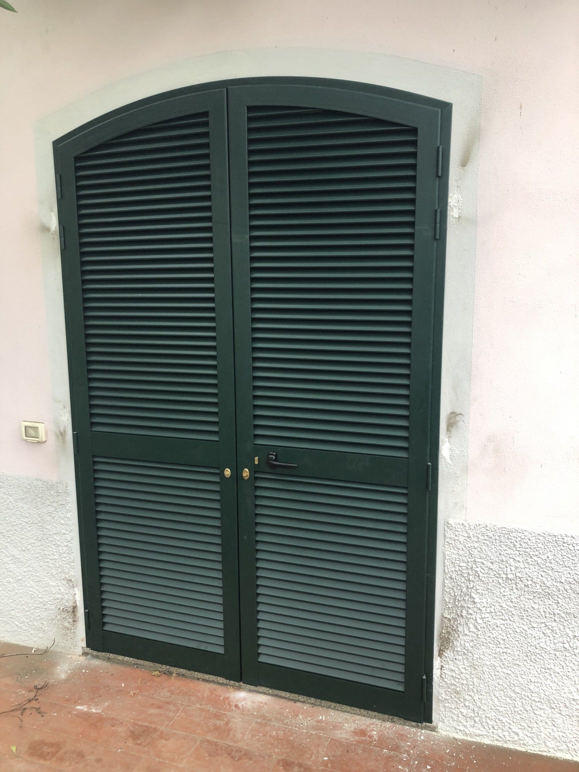 Doppie porte a persiana verde scuro con parte superiore arcuata su una parete rosa chiaro e strutturata.