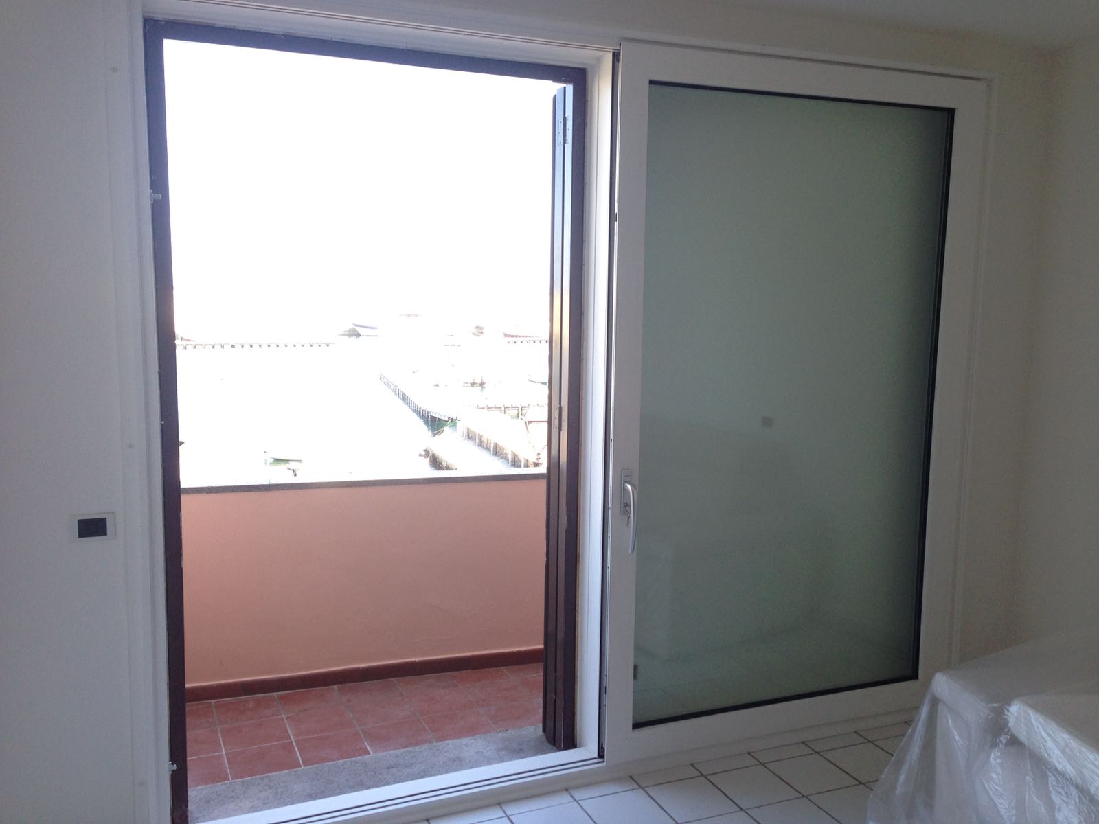 Porta scorrevole in vetro che si apre su un balcone; telaio bianco, pavimento in piastrelle marroni, vista esterna luminosa.