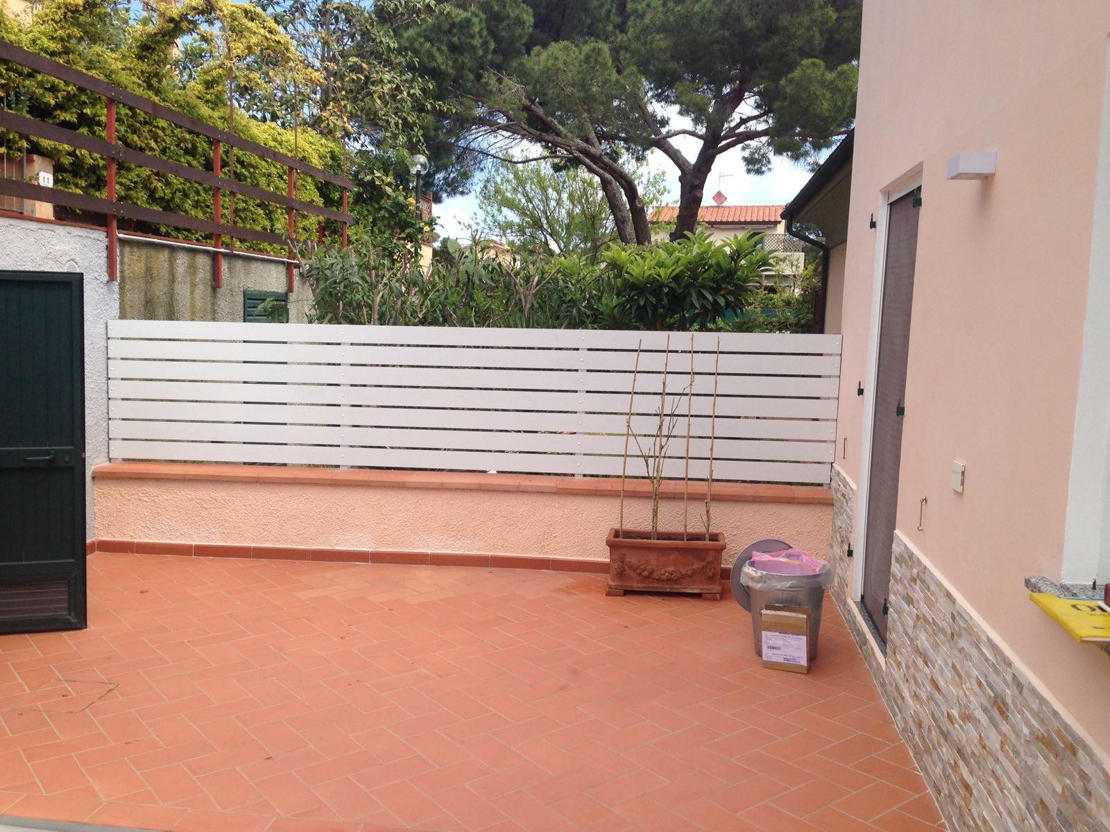 Terrazza con staccionata bianca, piastrelle in terracotta e un edificio rosa. Porta verde sulla sinistra.