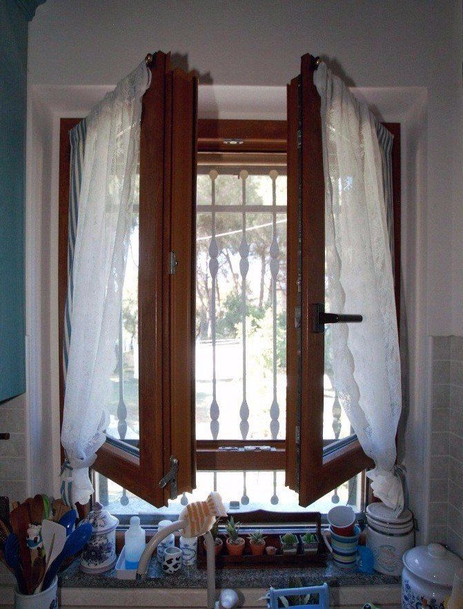 Finestra con due telai in legno aperti, tende bianche e inferriate di sicurezza, con vista sugli alberi.