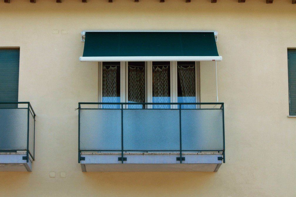 Tenda verde sopra una finestra con balcone in vetro. Esterno dell'edificio beige chiaro.