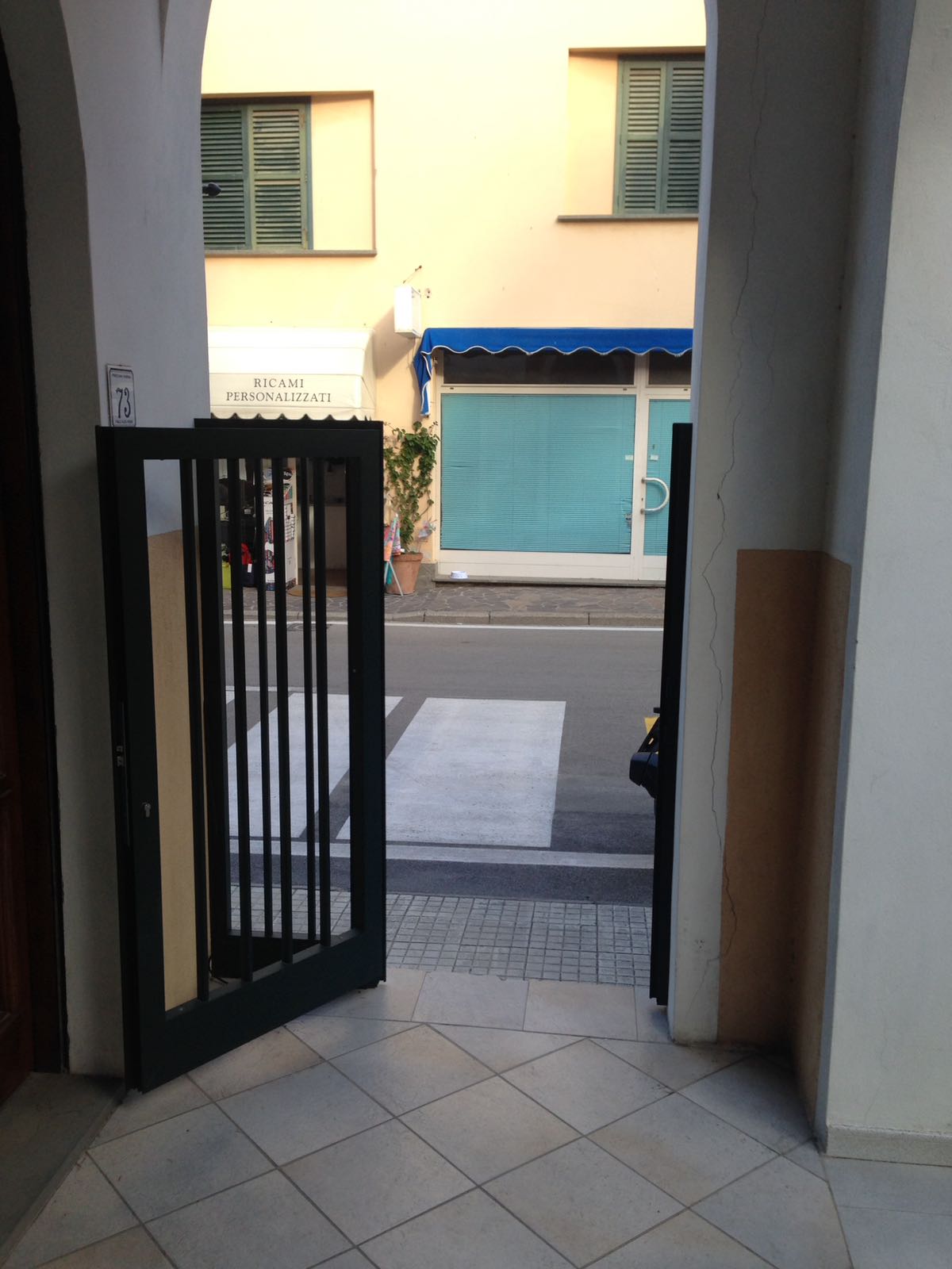 Cancello aperto in metallo nero che conduce a una strada con attraversamento pedonale e negozio chiuso. Edifici beige e bianchi lo circondano.