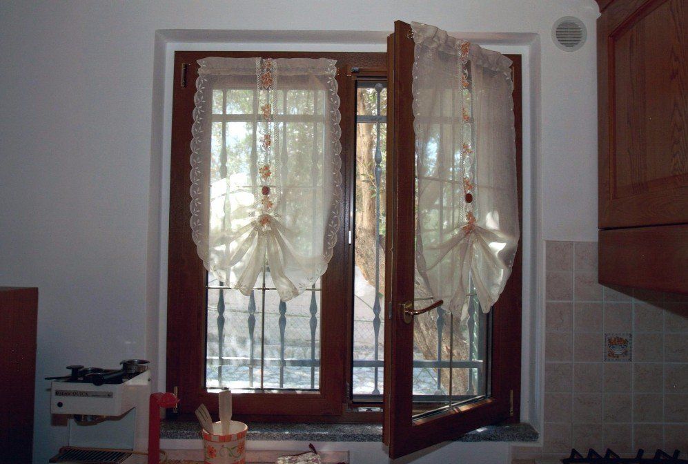 Finestra in legno aperta con tende trasparenti, vista sull'esterno. Ambiente cucina.