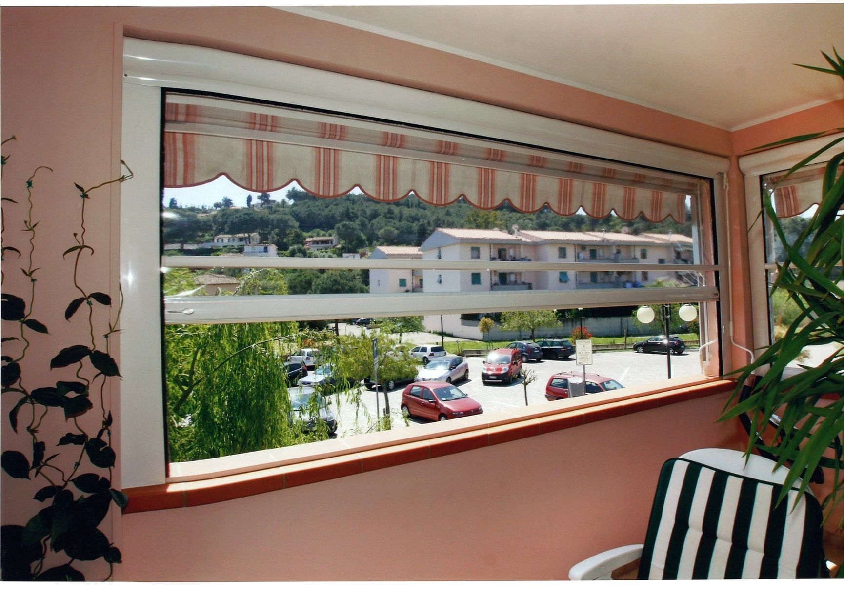 Vista dalla finestra: tenda a strisce, auto parcheggiate, edificio. Una sedia a strisce bianche e nere sulla destra.