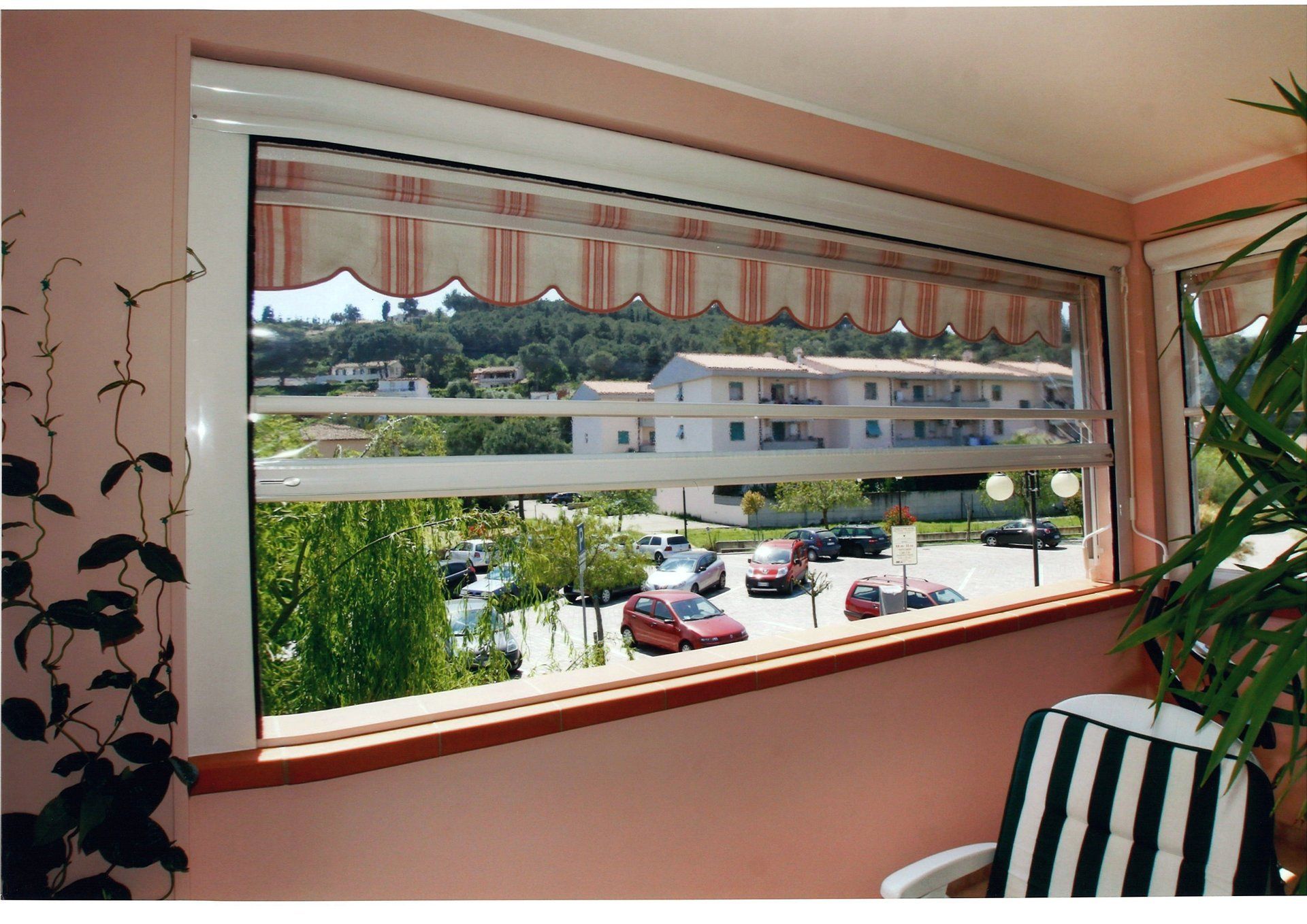 Vista dalla finestra: tenda a strisce, auto parcheggiate, edificio. Una sedia a strisce bianche e nere sulla destra.