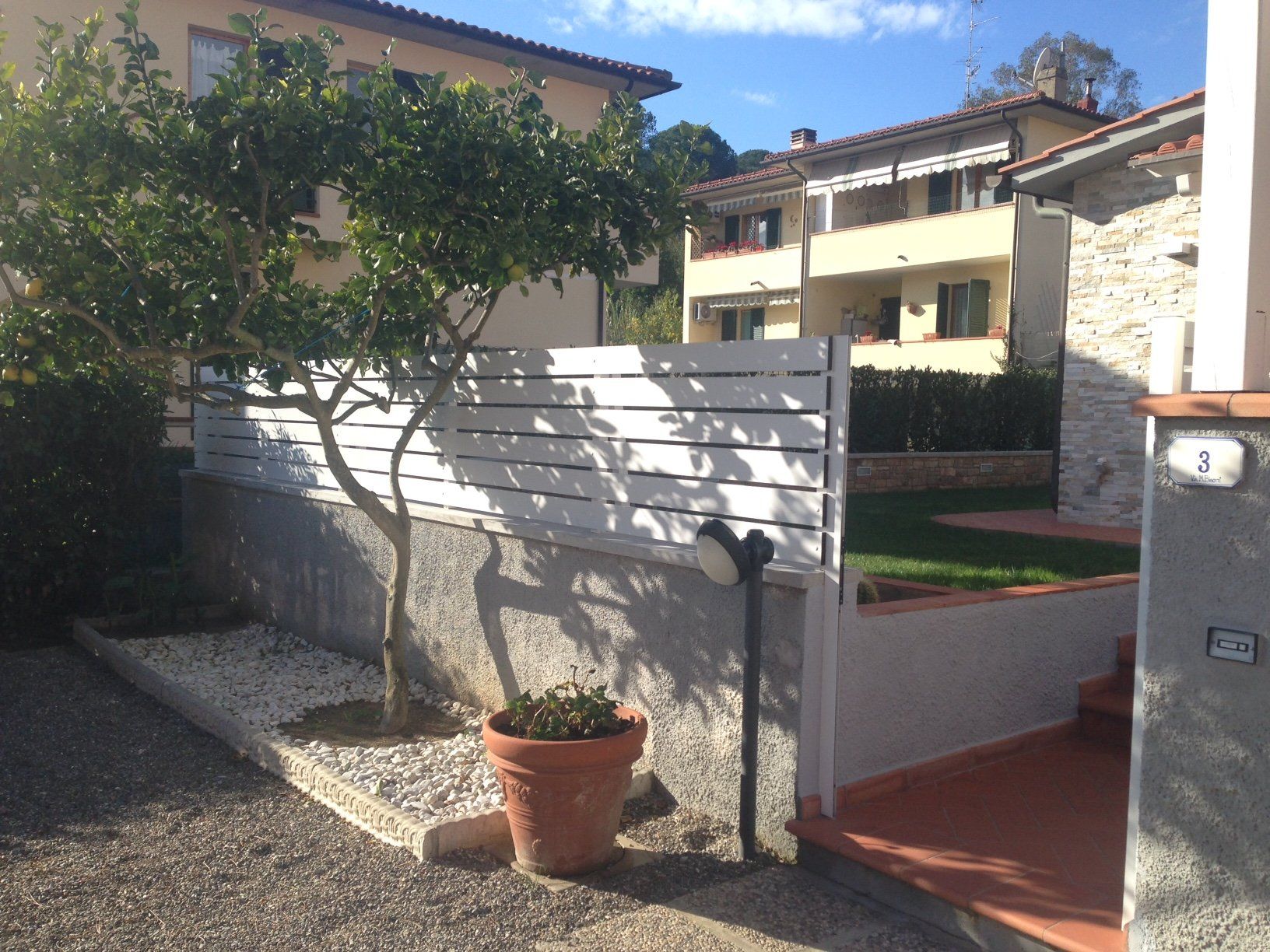 Un piccolo cortile con una staccionata bianca, un albero e un vaso di fiori di fronte a un edificio. Giornata di sole.