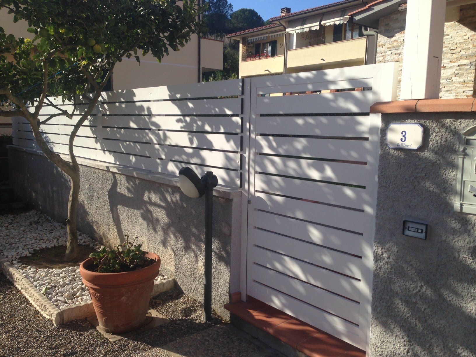 Recinzione e cancello in doghe bianche davanti a una casa. Nella scena sono presenti un albero e una pianta in vaso.