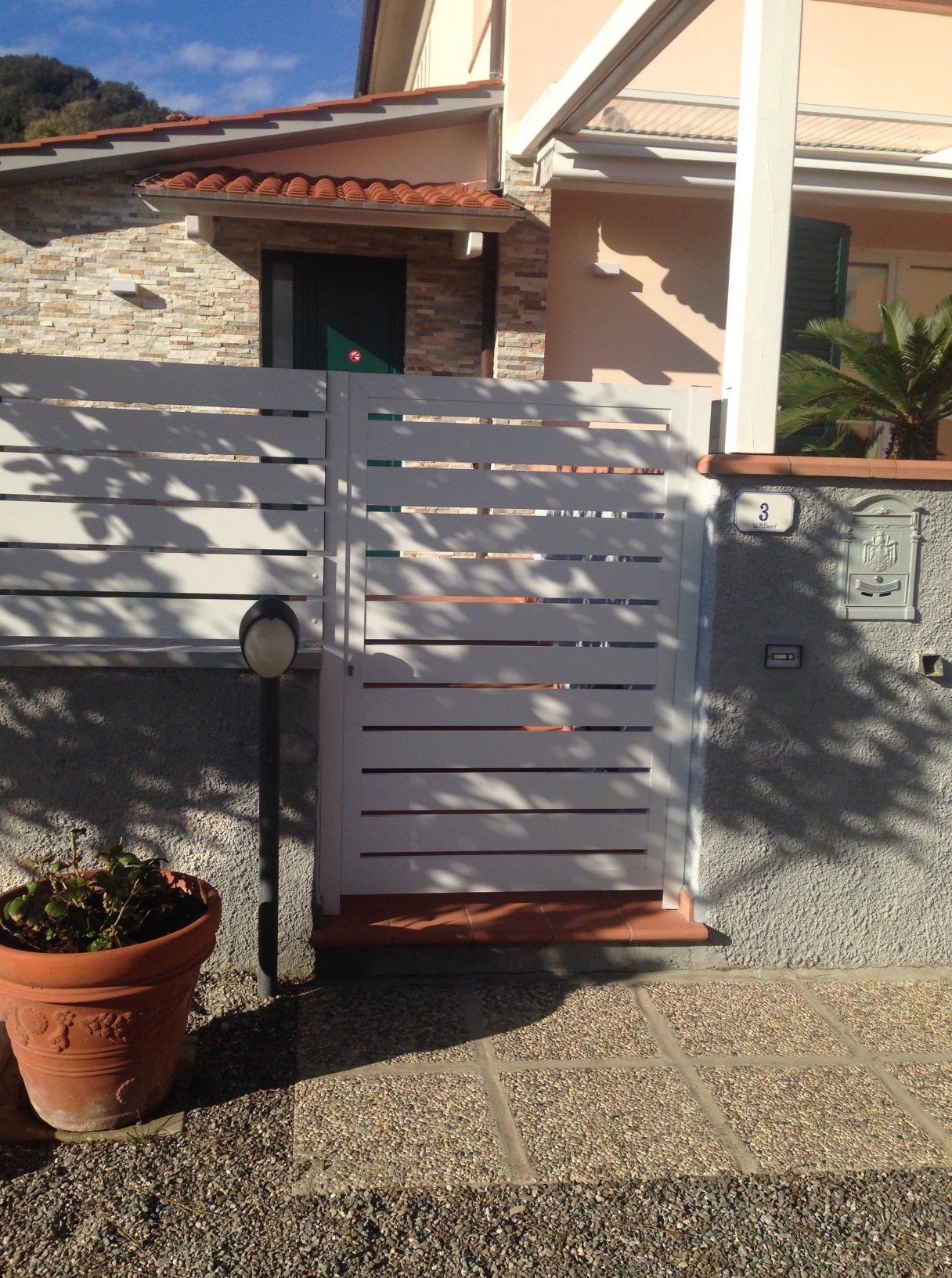 Cancello bianco con stecche davanti a una casa, con una pianta in vaso sulla sinistra e un vialetto in pietra.