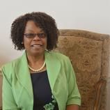 A woman is sitting in a chair wearing a green jacket and glasses.