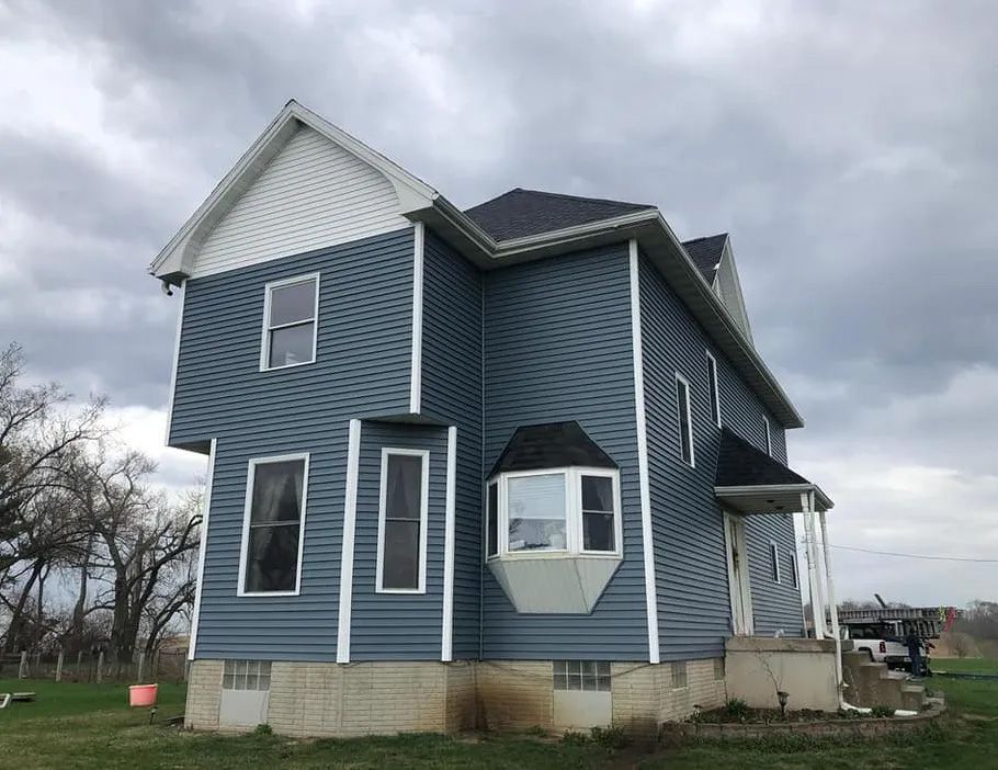 A large blue house with white siding and a black roof is sitting on top of a lush green field.