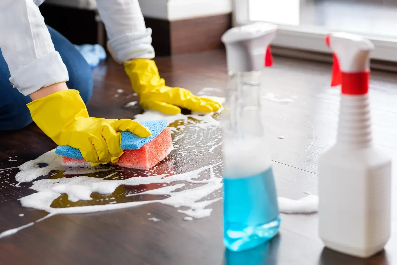 Person wearing yellow gloves cleans floor with sponge and spray bottles.