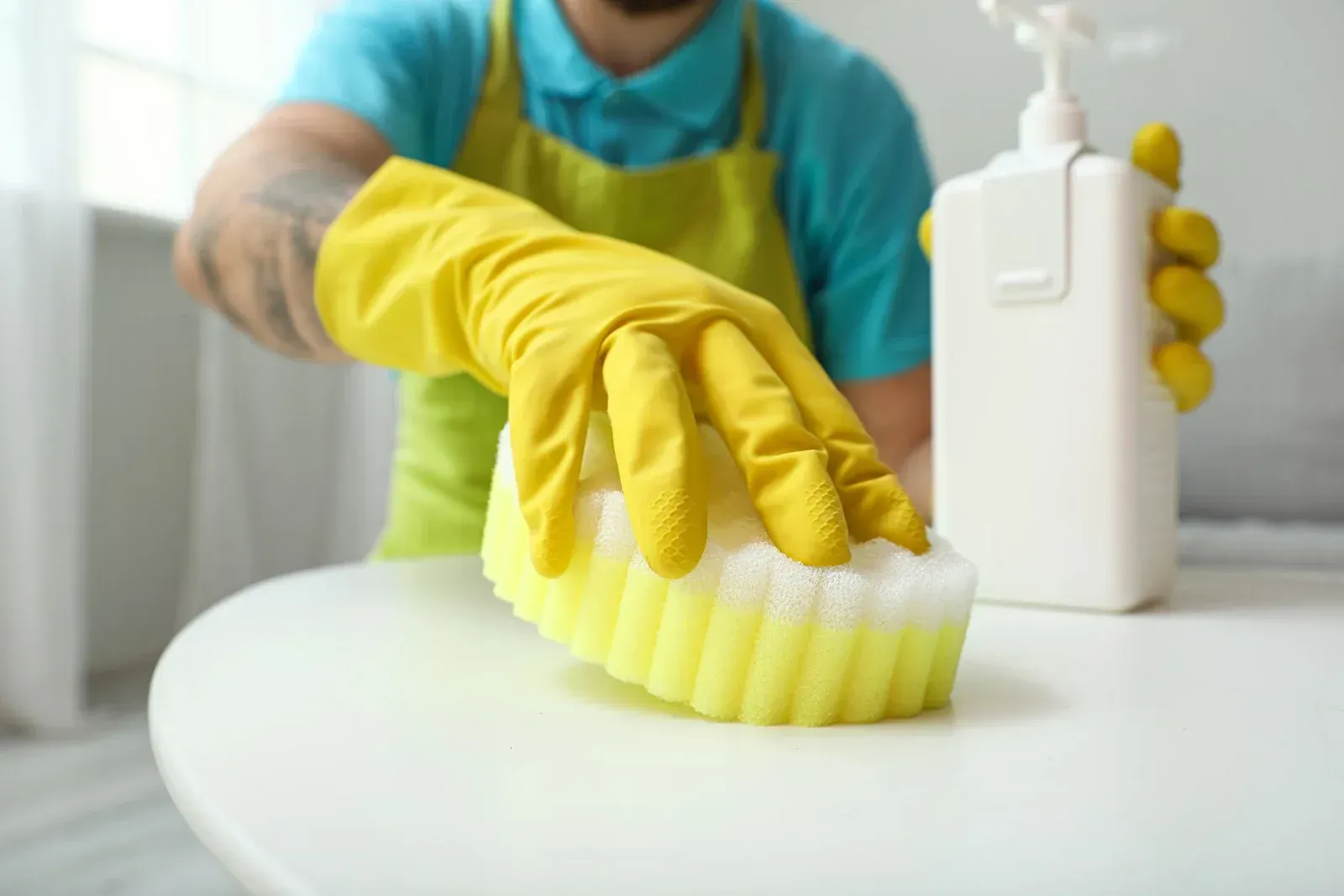 Person wearing yellow gloves cleans a white table with a sponge and spray bottle.