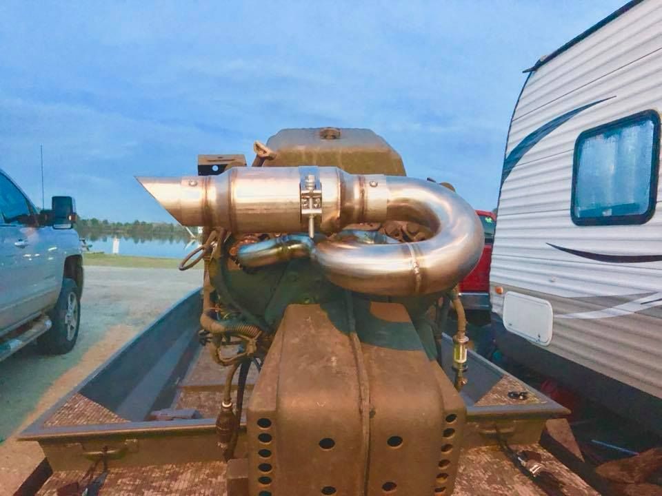 Mud boat engine with curved exhaust, in a metal boat, next to a lake and parked vehicles.