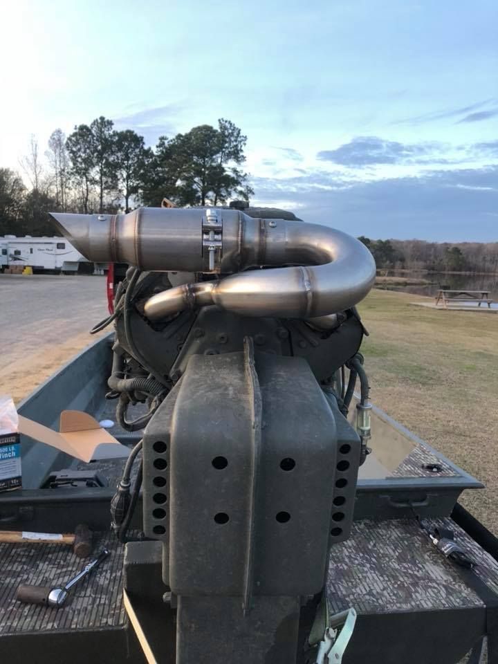 Boat motor with curved stainless steel exhaust pipe. Mounted on a green boat, outdoor setting.