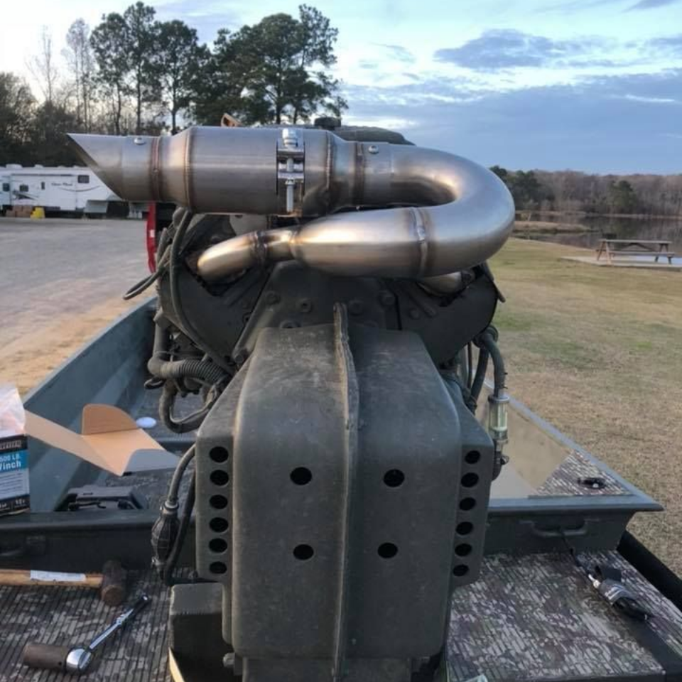 A mud boat engine with a custom exhaust pipe in a grassy outdoor setting, near a body of water.