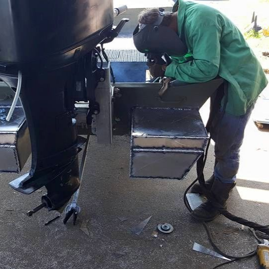 Welder in green jacket welds metal box on a boat's outboard motor.