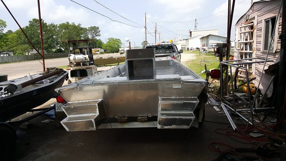 Aluminum boat under construction in a workshop. Rear view shows stepped hull, storage box.
