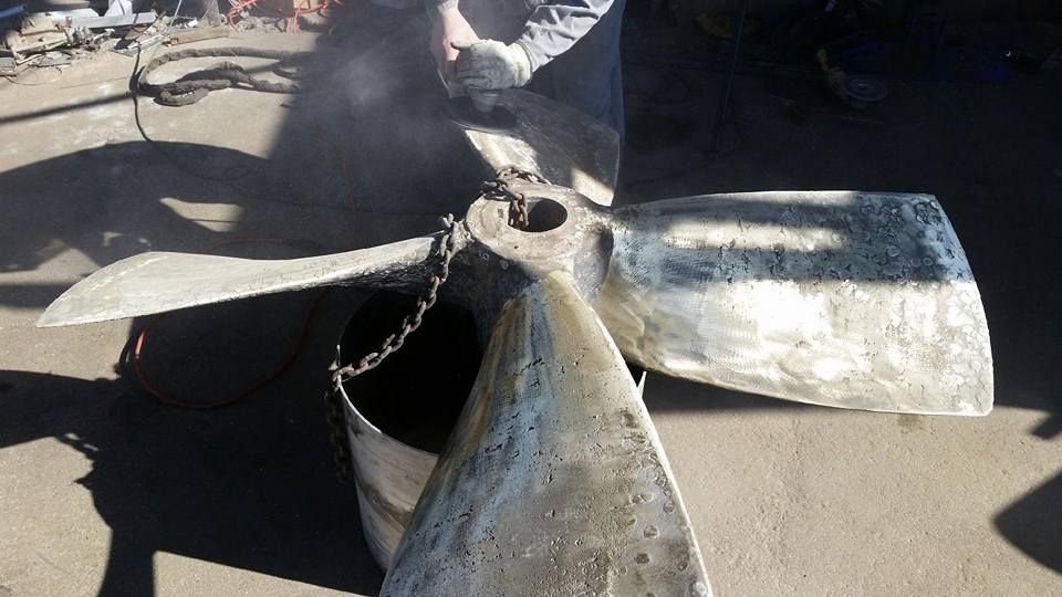 Large, worn metal propeller on a concrete surface, with a chain and bucket visible.