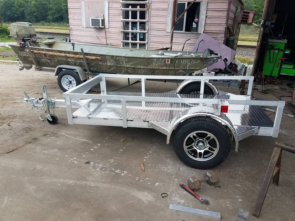 Aluminum utility trailer with a boat in the background, parked near a building.