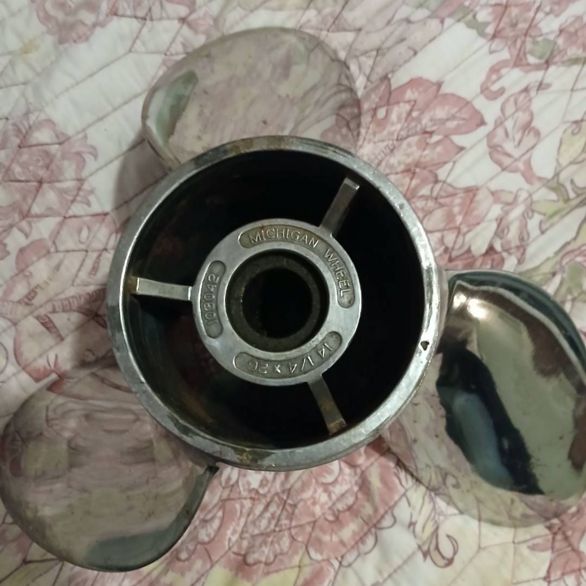 A stainless steel boat propeller with three curved blades, sitting on a floral patterned surface.