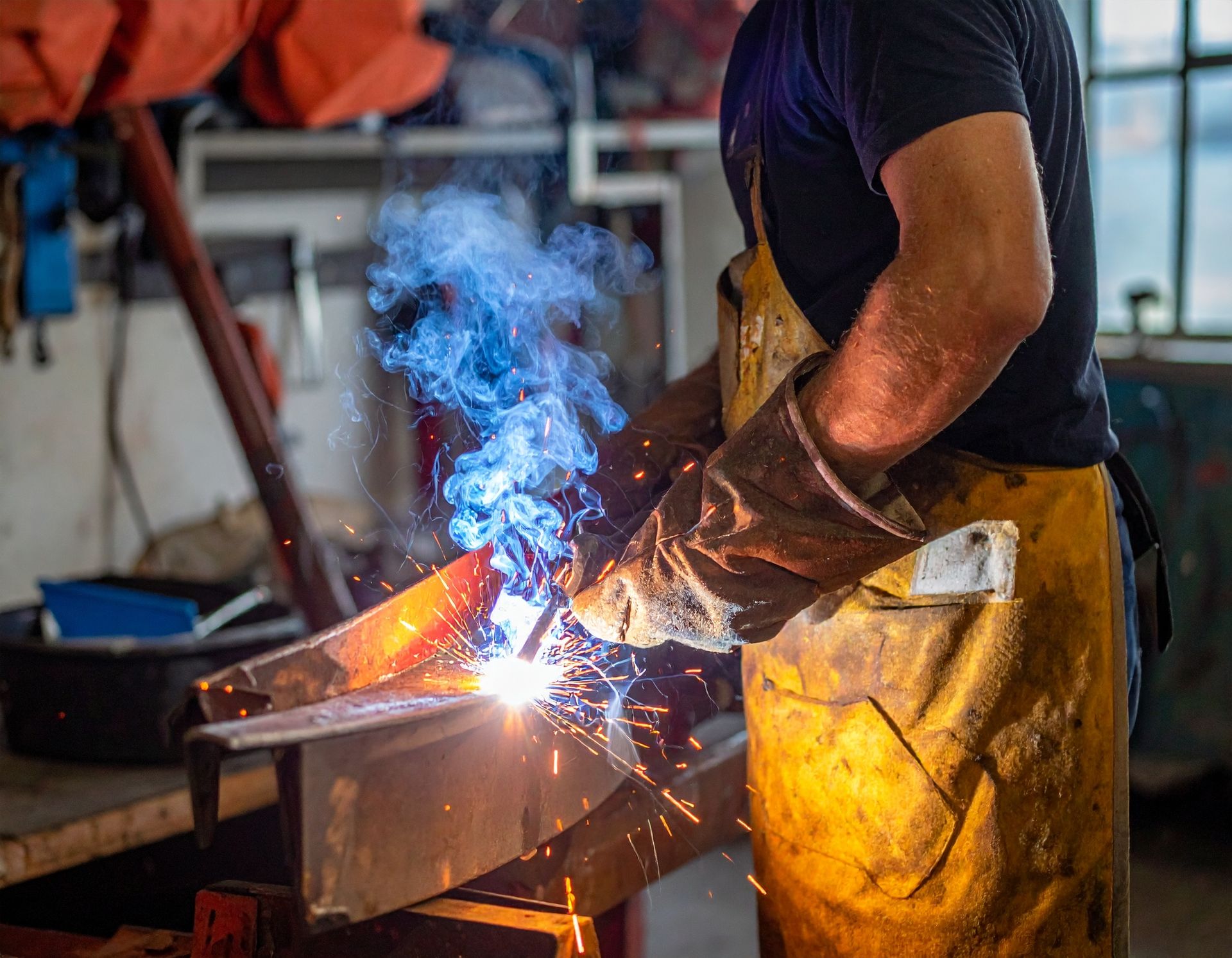 Welder in workshop, wearing gloves and apron, sparks flying from metal being worked on.