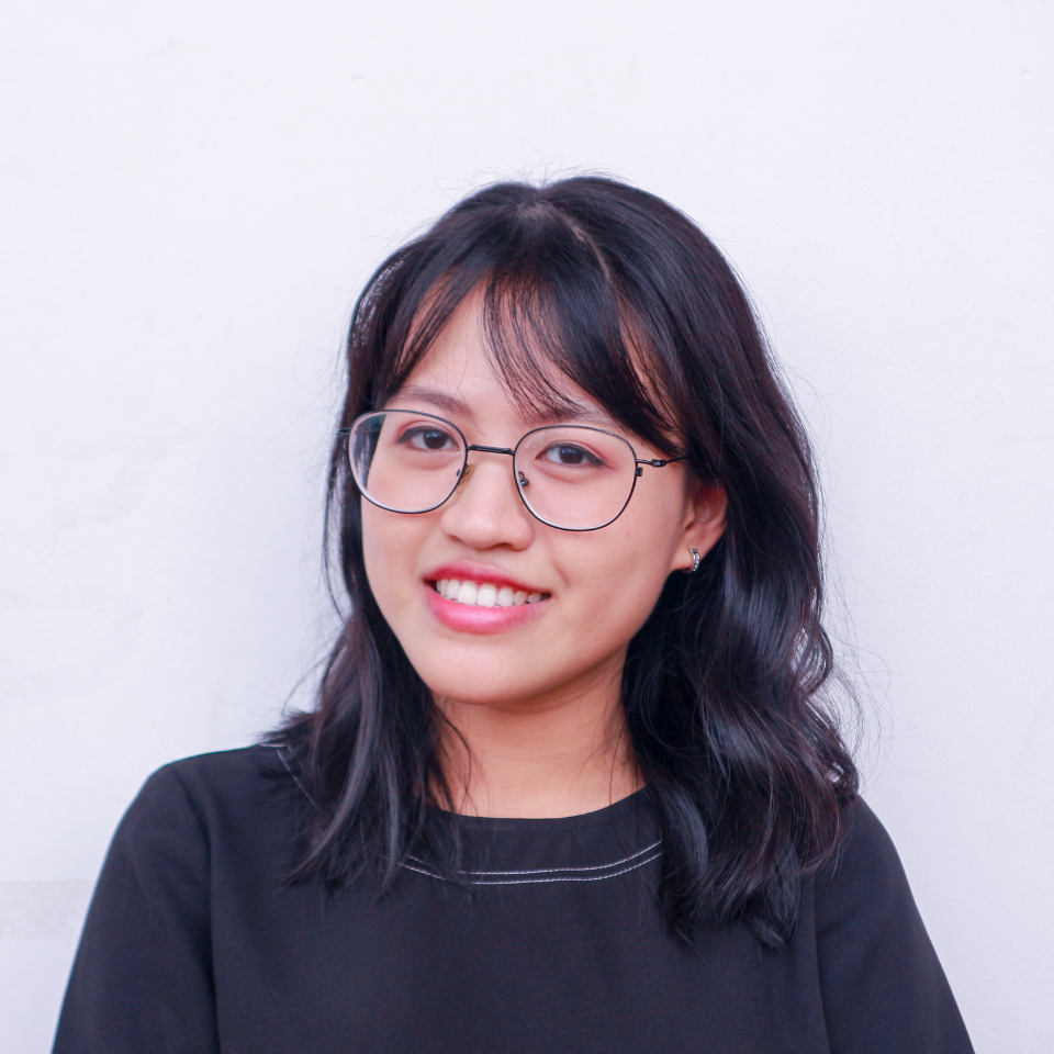 Woman with glasses smiles, black hair, dark top, against a white background.