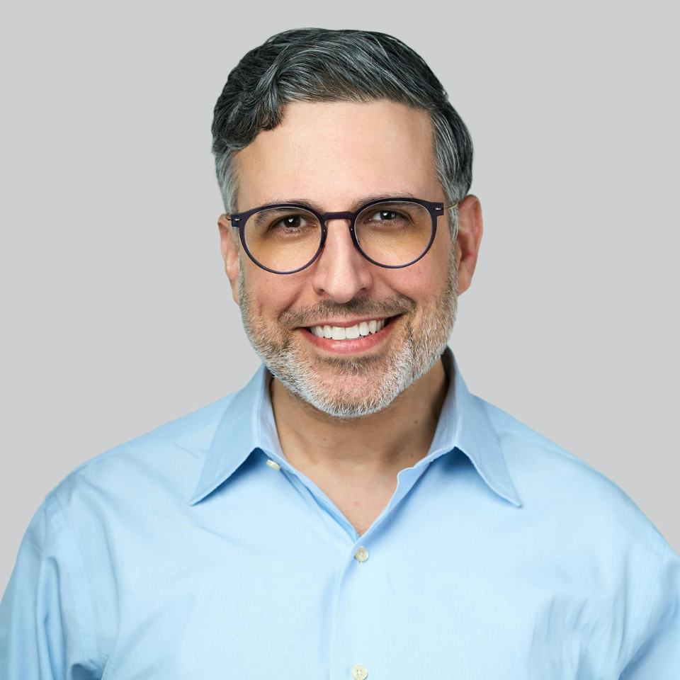 Man with glasses, light blue shirt, smiling. Gray background.