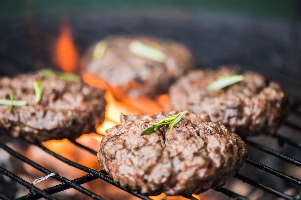 Grilled burger patties with herbs on a hot grill, flames visible.