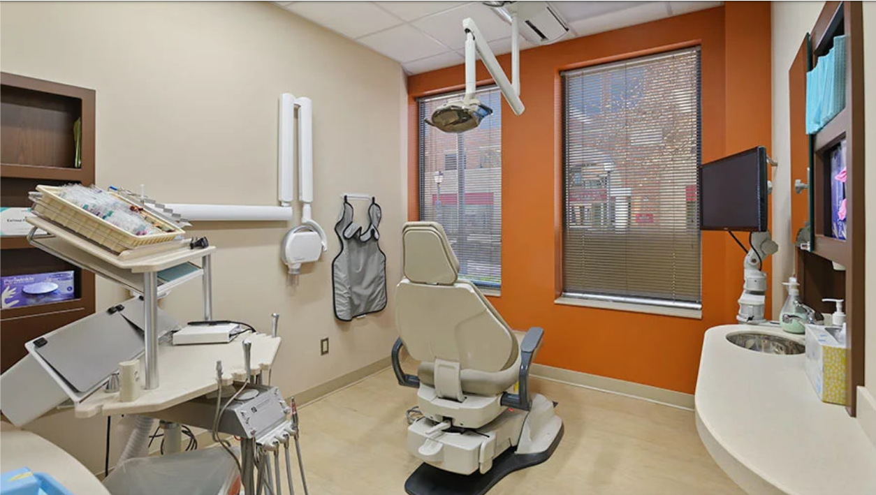 Dental operatory with chair, equipment, and window, with an orange accent wall.