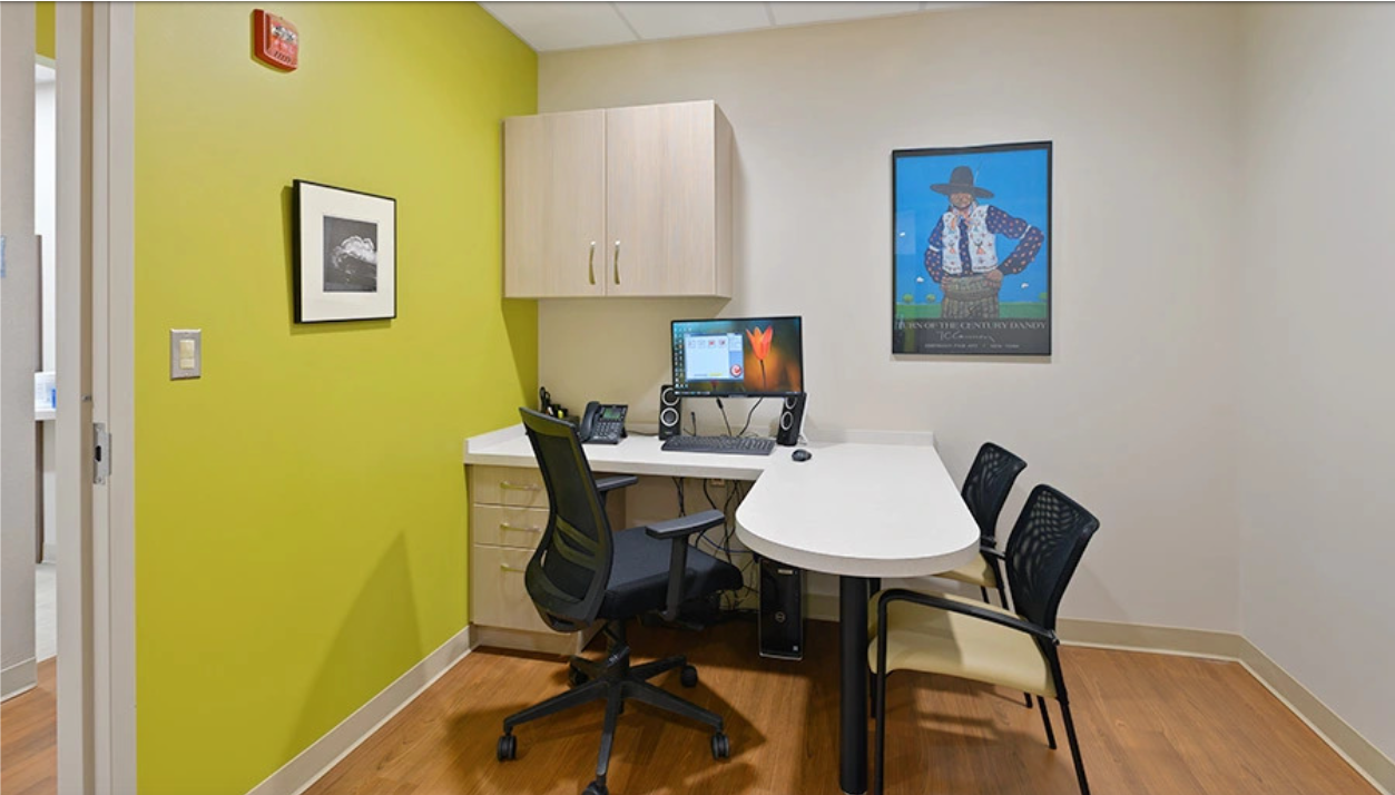 Examination room with desk, chairs, computer, artwork, and cabinets; lime green wall.