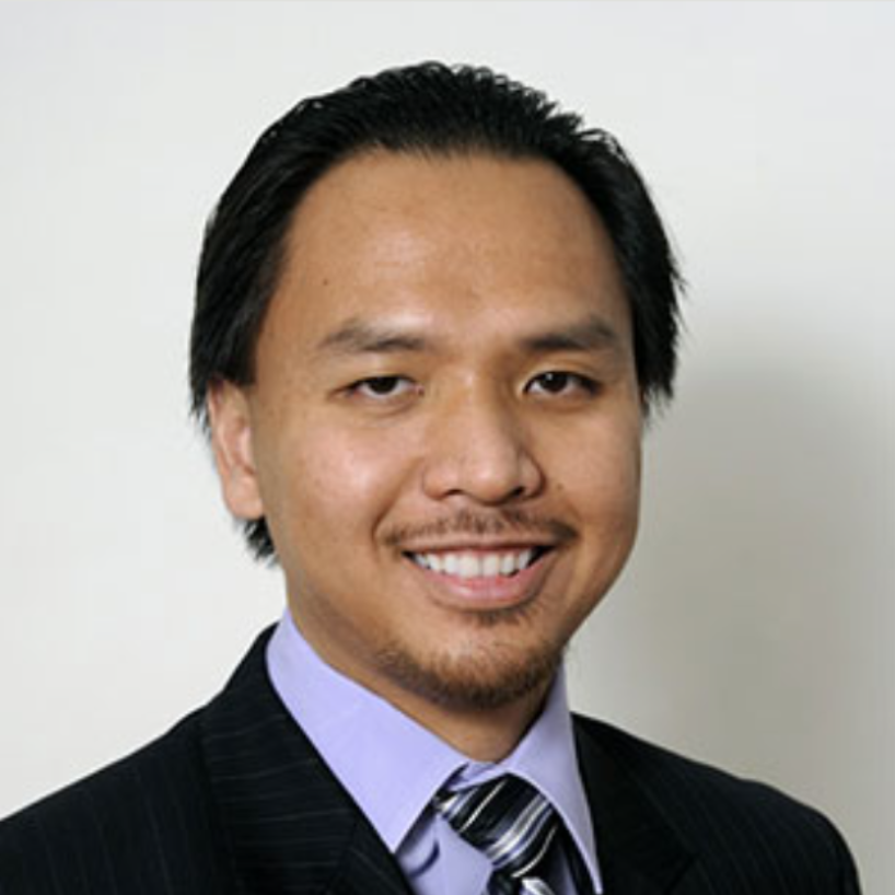 Man in suit smiles at the camera, light skin, short dark hair, blue shirt, dark tie, white background.