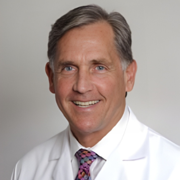 Man in a white coat and patterned tie smiles; studio portrait.