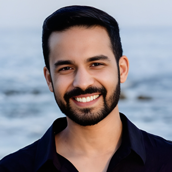 Man with dark hair and beard smiles broadly outdoors, ocean in the background.